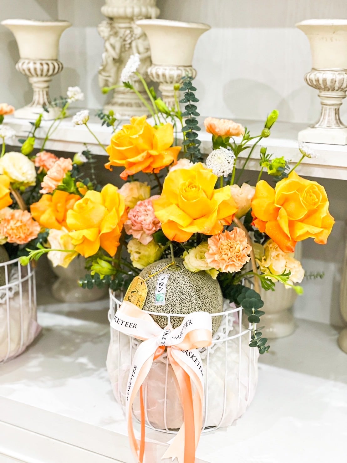 A decorative arrangement of orange roses, melon fruit, and yellow flowers is displayed in a white wire basket, with a cantaloupe wrapped in a ribbon inside. The basket sits on a white shelf surrounded by ornate, white candle holders in the background.
