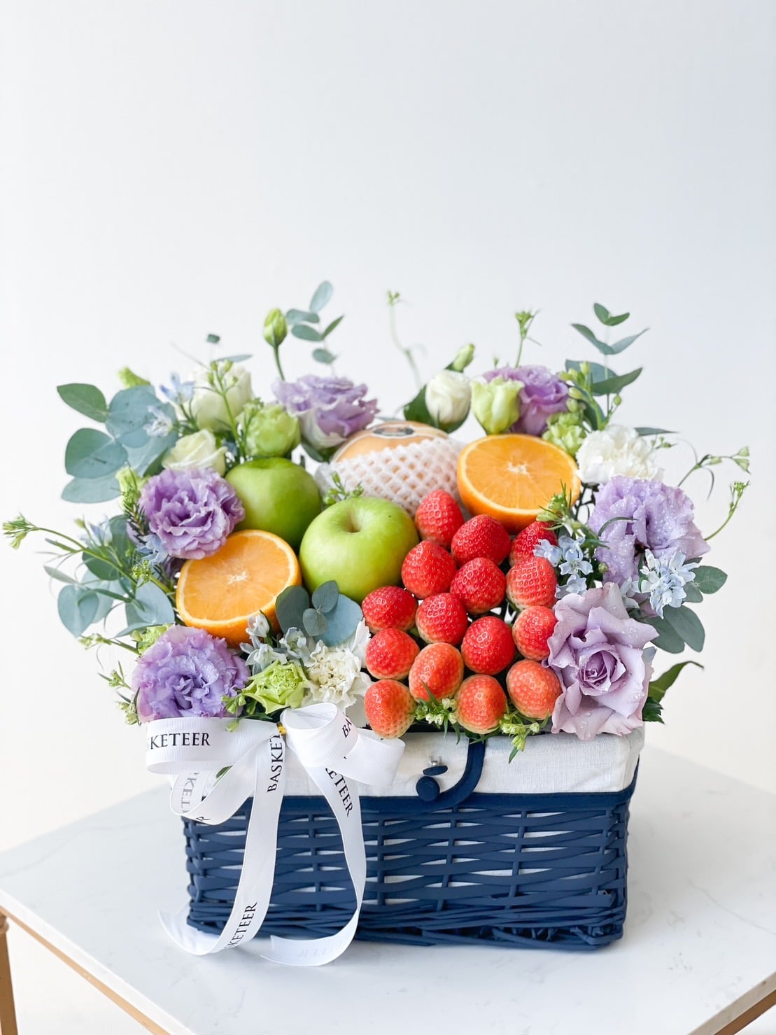 Introducing the Purple Flowers Deluxe Fruit Basket: A blue wicker basket filled with green apples, strawberries, a pomelo, and orange halves. Nestled among the fruits are purple and white flowers, green leaves, and a white ribbon with text. The background is plain.