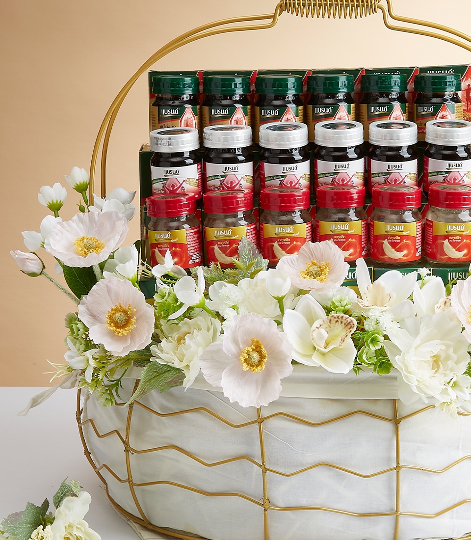 A luxurious Basketeer gift basket brimming with an exquisite selection of green and red jars, artfully arranged in two perfect rows. The basket is exquisitely adorned with delicate white and pale pink artificial flowers amidst lush greenery, creating an enchanting visual appeal. Its gold-colored handle and frame exude opulence, enhancing its sophisticated charm. A satin ribbon with the Basketeer logo elegantly drapes across the arrangement, completing this inviting masterpiece.