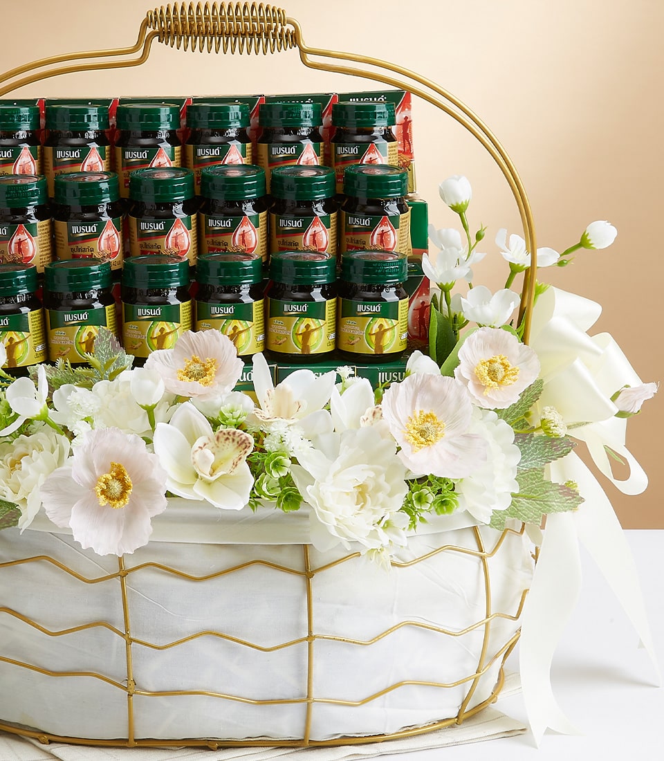 A luxurious Basketeer gift basket elegantly showcases an array of green glass jars, each adorned with vibrant red and yellow labels. The artfully arranged jars are neatly stacked in rows within the basket, which is beautifully embellished with an opulent arrangement of white roses and lilies. A large, sophisticated white ribbon, featuring the prestigious Basketeer logo, gracefully accentuates the side of this captivating display.