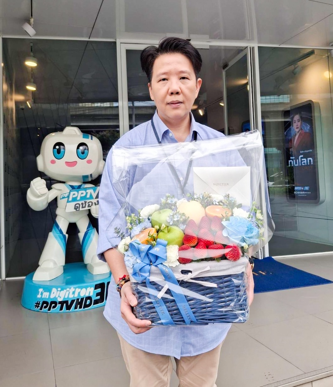 A person holding a Purple Flowers Deluxe Fruit Basket, adorned with blue and white ribbons, stands in front of a building with glass doors. The basket overflows with fruits and flowers. A robot mascot and a poster are visible in the background.