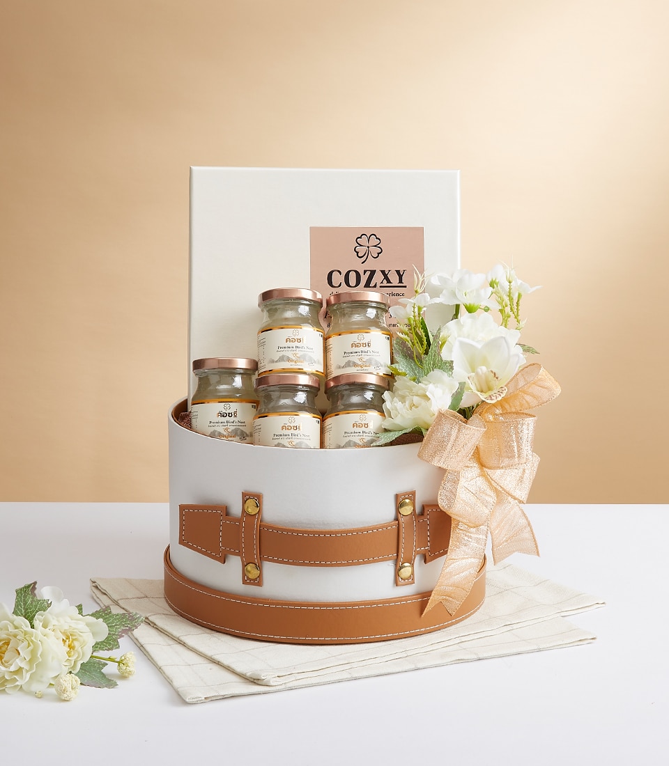 A luxurious gift box with an elegant tan leather strap, featuring six exquisitely packaged jars of gourmet delights. A beautiful bouquet of white flowers is gracefully tied with a peach ribbon, adding a touch of sophistication. The pristine white box behind the jars proudly displays the 
