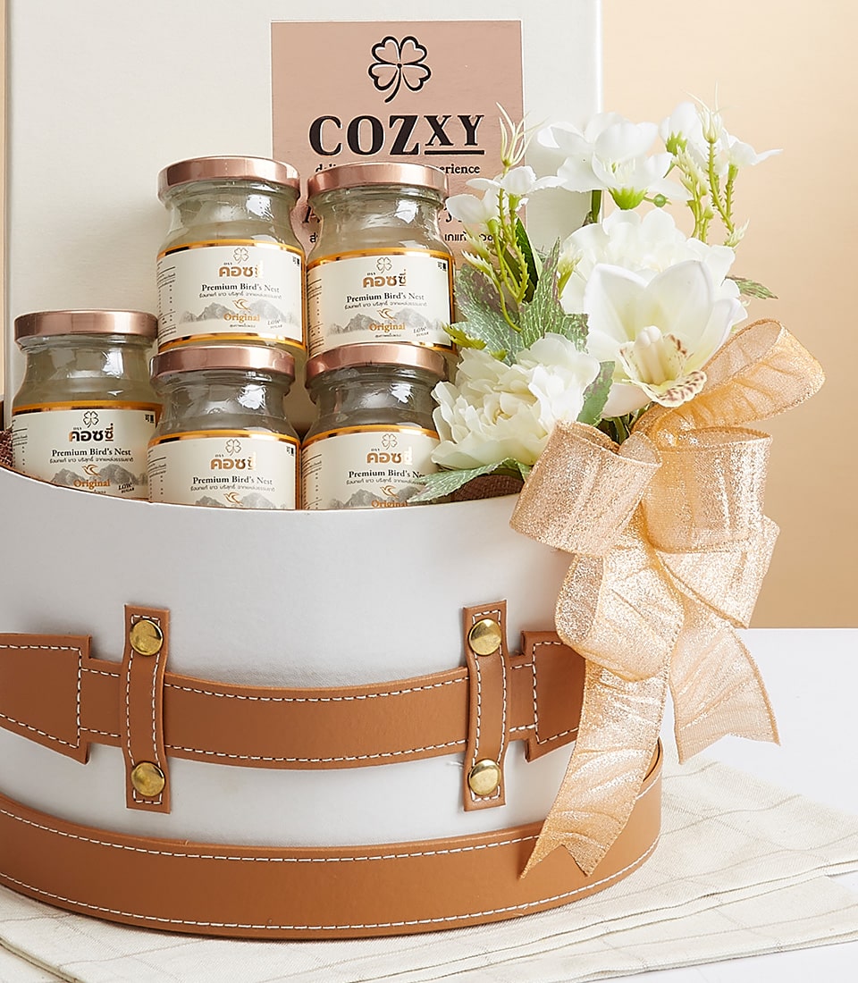An exquisite Basketeer gift box, elegantly adorned in white and beige tones, houses four premium glass jars with sophisticated yellow labels. Each jar is crowned with delicate clusters of pristine white flowers and a luxurious gold ribbon. A stylish card emblazoned with 