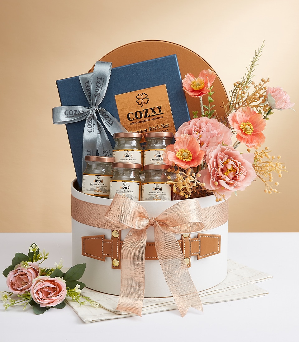 A round, opulent white Basketeer gift basket graced with a lavish pink ribbon. Inside, discover a sophisticated blue box tied with an elegant silver ribbon and nestled among several artisanal jars bearing refined beige labels. Abundant faux pink blooms accentuate the arrangement, while additional delicate pink flowers are artfully scattered on a pristine white surface, completing this luxurious display.