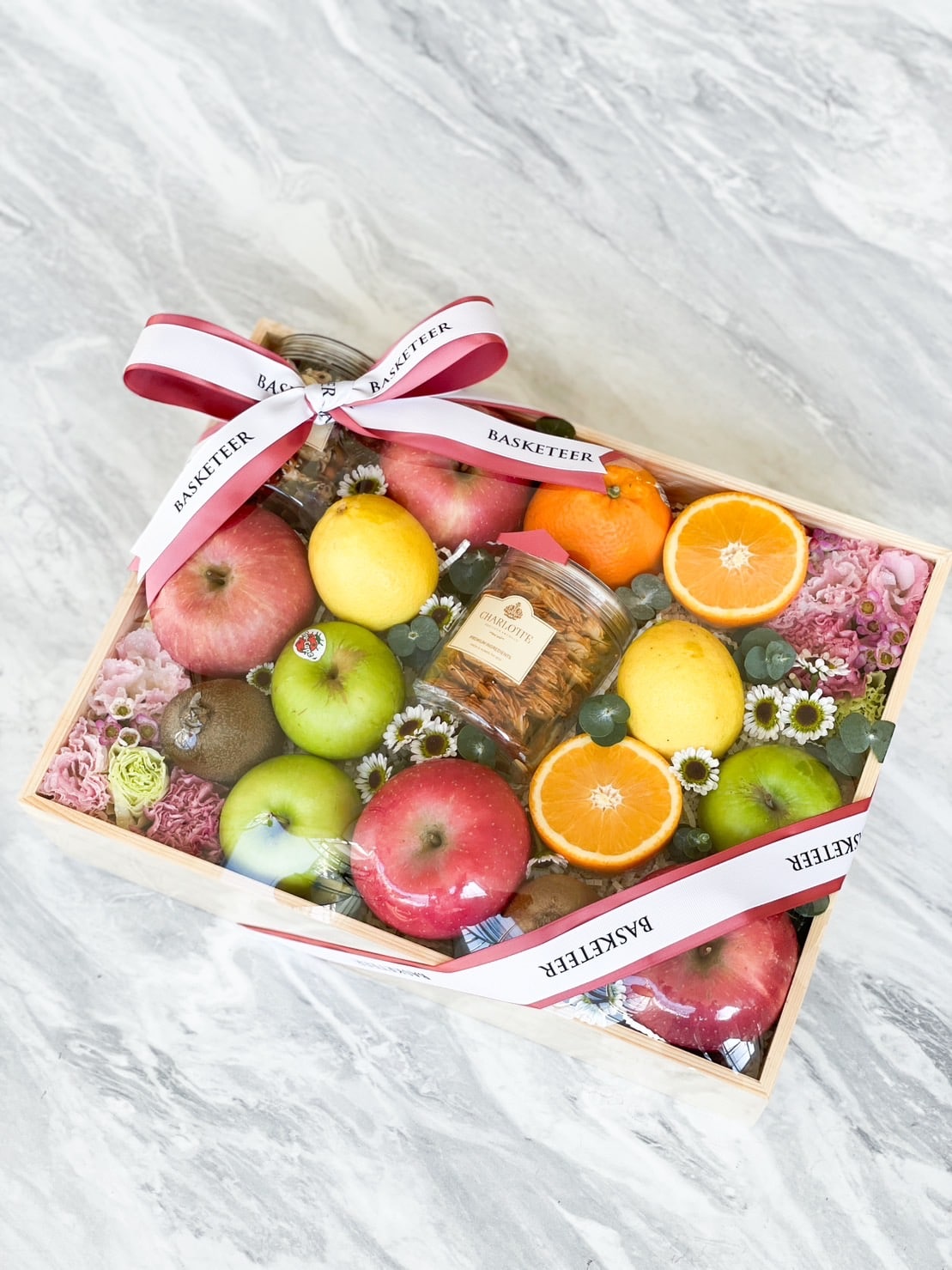 An Exotic Fruit Gift Box filled with assorted fresh fruits, including apples, oranges, lemons, grapes, and figs. The wooden box also contains a jar of mixed nuts and is decorated with a ribbon and small white flowers, all set on a marble surface.