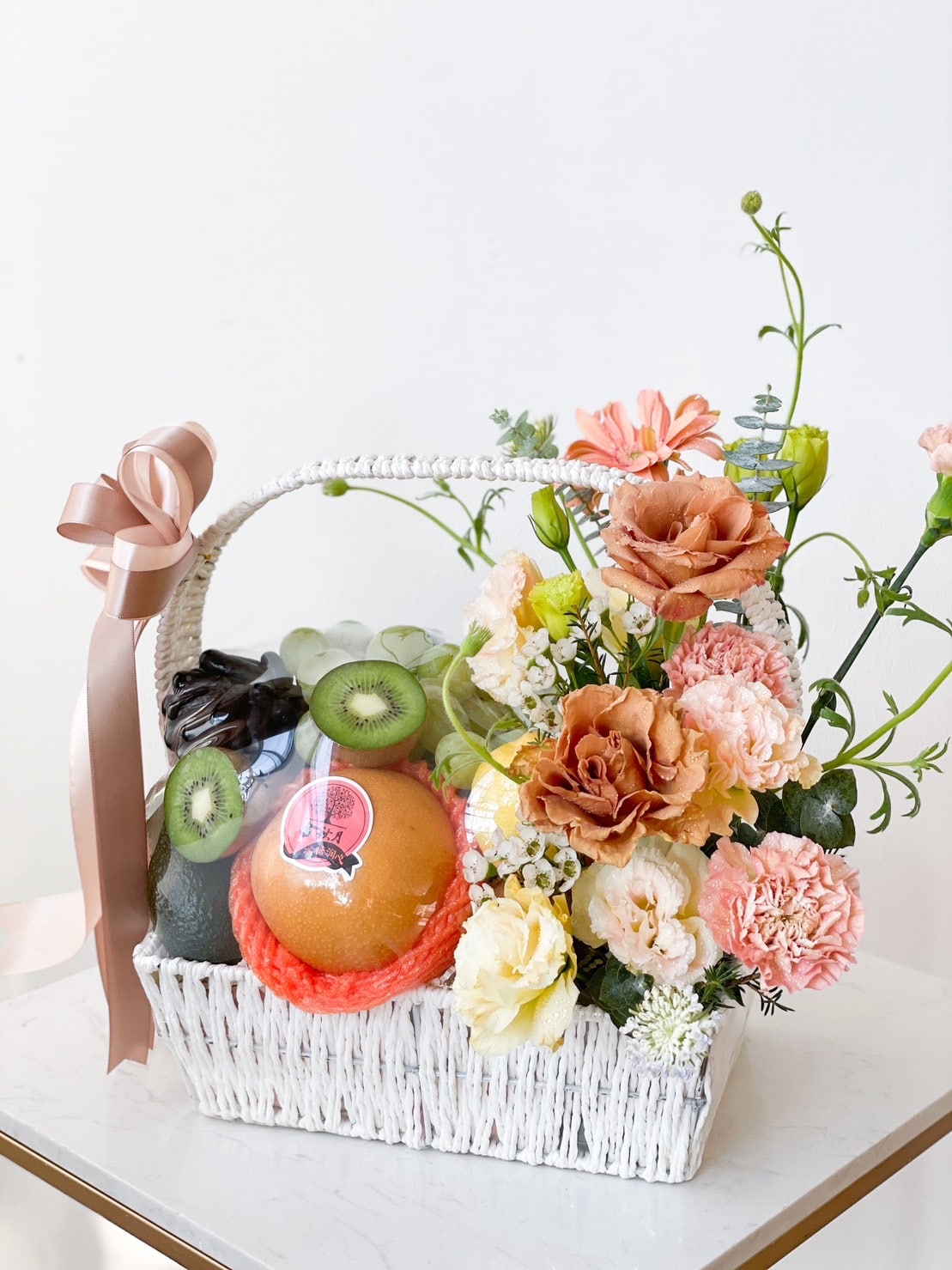 A white wicker basket decorated with a peach ribbon holds an arrangement of various fruits, including a large pomelo, kiwis, and apples, alongside a colorful assortment of flowers such as roses, carnations, and green sprigs. This delightful Blue Rose Fruit Basket adds charm to any setting.