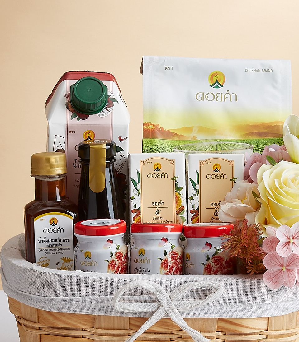 A luxurious wicker basket, exquisitely presented, brims with an assortment of premium Doi Kham brand products. Nestled amid delicate white and pink flowers, the selection includes elegantly boxed items, bottled liquids in glass containers, artisanal jars, and a pristine carton. Each product proudly displays the distinctive Doi Kham logo and intricate Thai script labels. The entire arrangement exudes sophistication with a ribbon or box adorned with the prestigious Basketeer emblem, elevating the gift to a truly inviting experience.