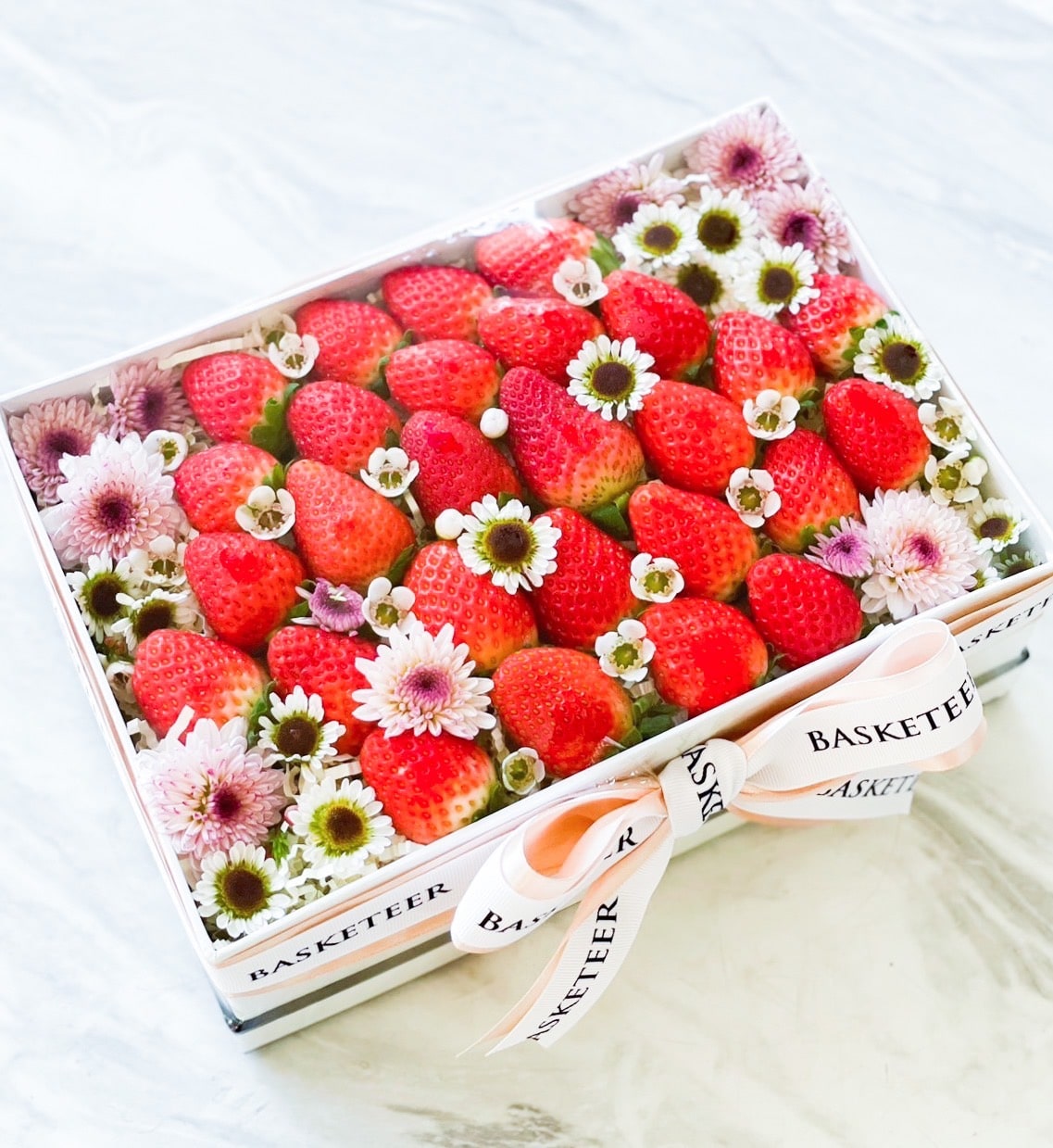 A Strawberry Bliss Gift Box filled with fresh strawberries and interspersed with small pink and white flowers. The box is adorned with a white ribbon that has 