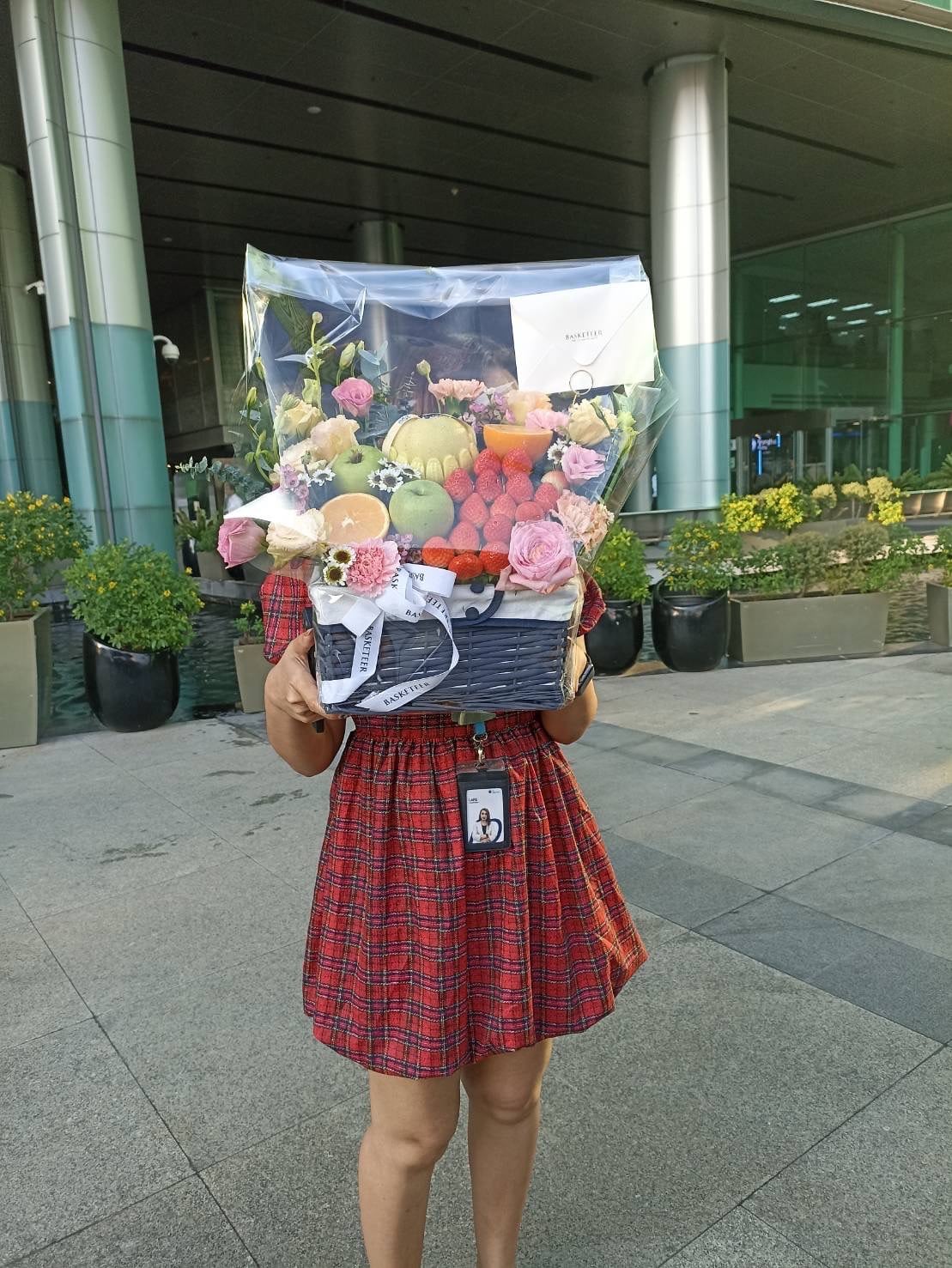 A person in a red plaid dress holds a Deluxe Fruit Basket brimming with assorted fruits and purple flowers. They are standing outside a building with large glass windows and several green plants in the background. The person's face is hidden behind the vibrant basket.
