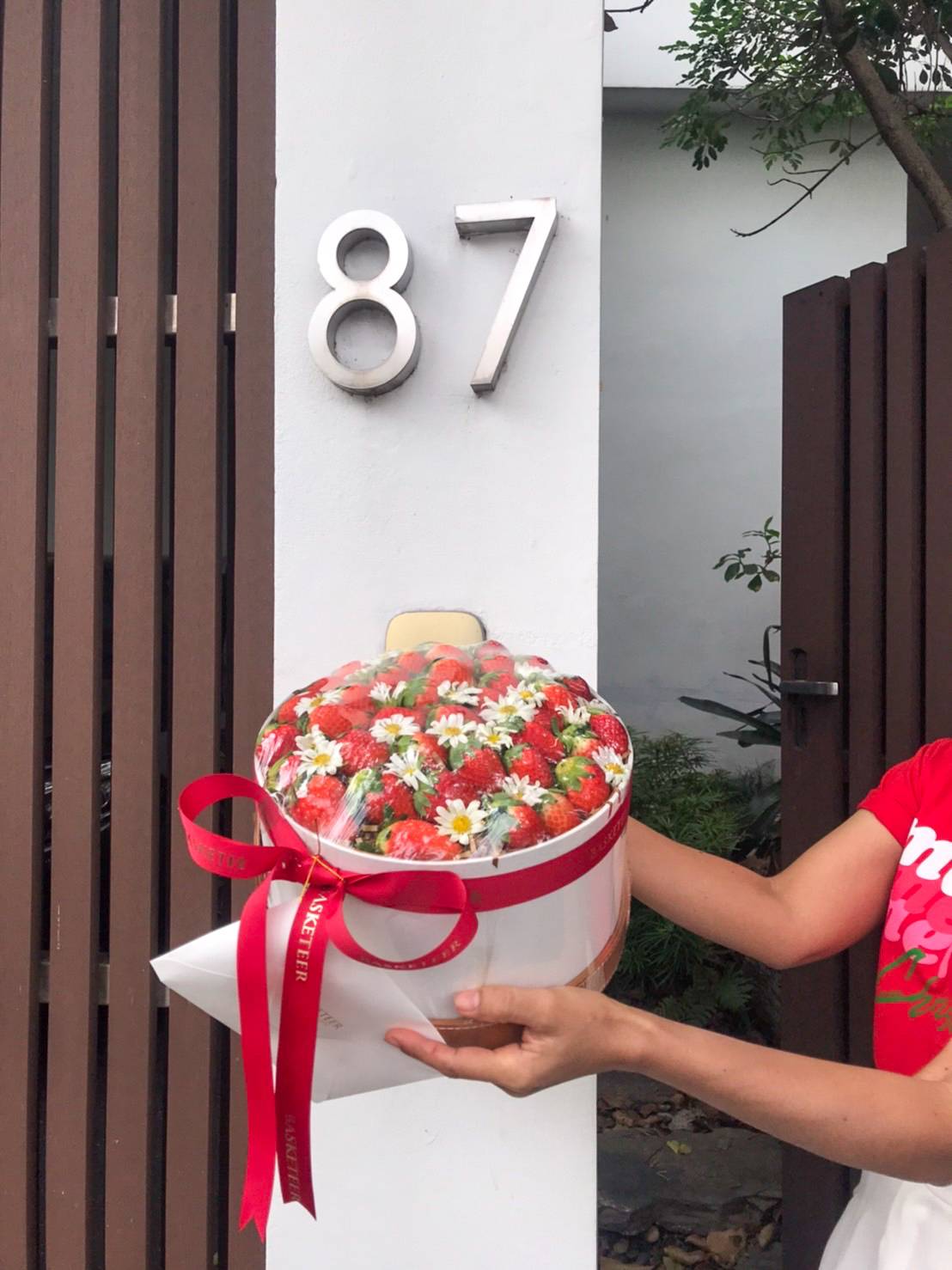 A person in a red shirt is holding a bouquet of strawberries and flowers artfully wrapped in white paper with a red ribbon, standing outside a house numbered 87. It almost looks like they are presenting an elegant strawberry gift box.