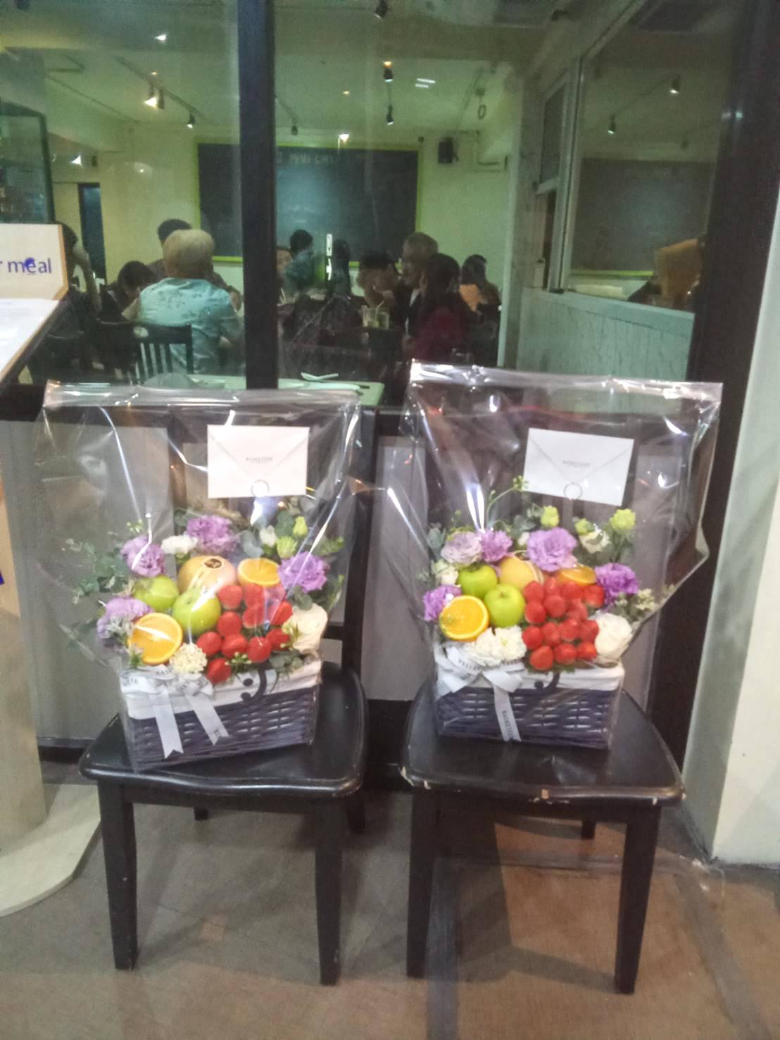 Two black chairs outside a glass-walled cafe, each holding a Purple Flowers Deluxe Fruit Basket filled with various fruits like oranges, apples, and strawberries. The baskets are wrapped in clear plastic and adorned with white ribbons, showcasing an assortment of white and purple flowers.