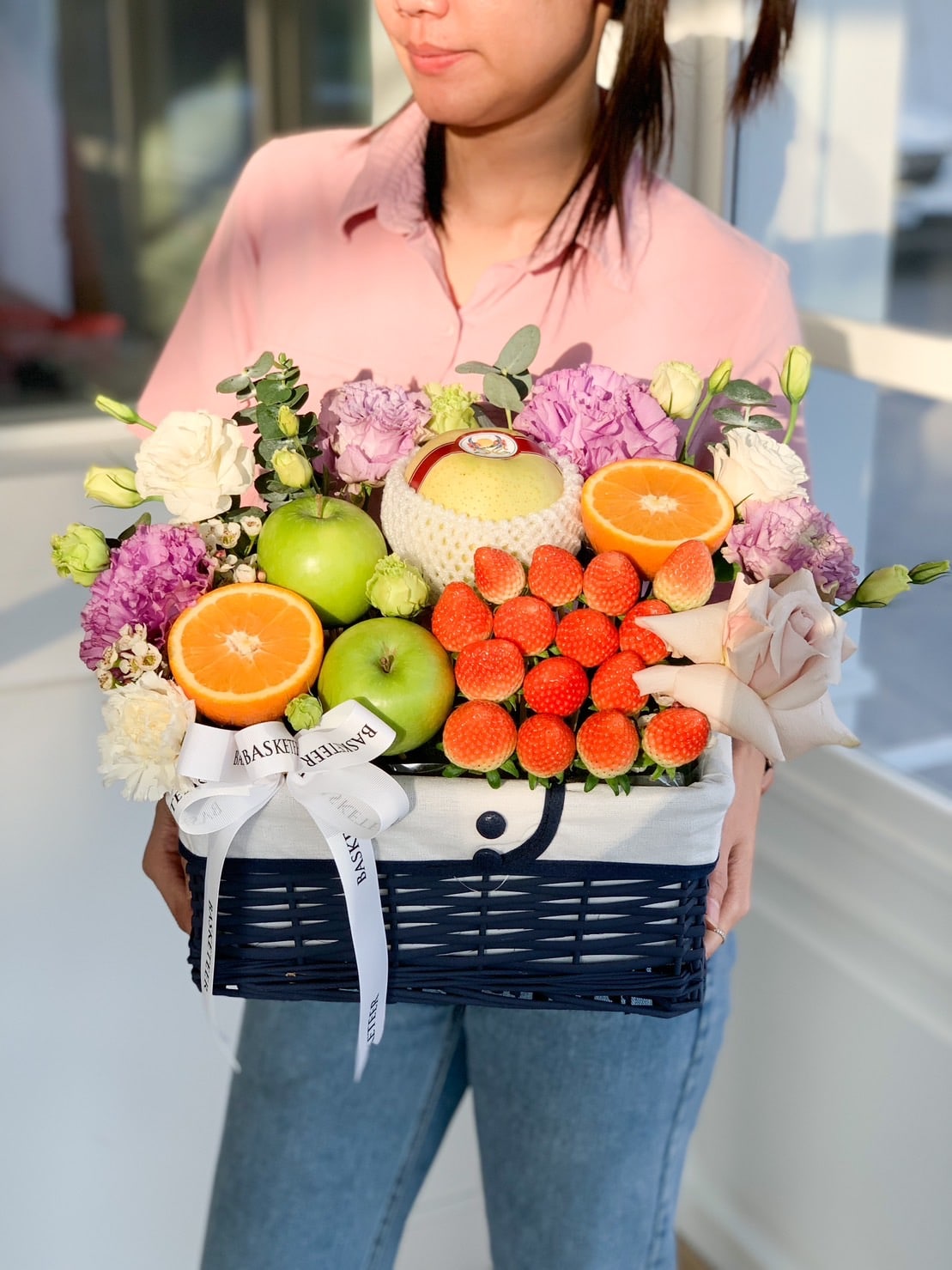 A person wearing a pink shirt holds a Purple Flowers Deluxe Fruit Basket filled with green apples, strawberries, oranges, a wrapped fruit, and vibrant purple and white flowers. A white ribbon is elegantly tied to the handle.