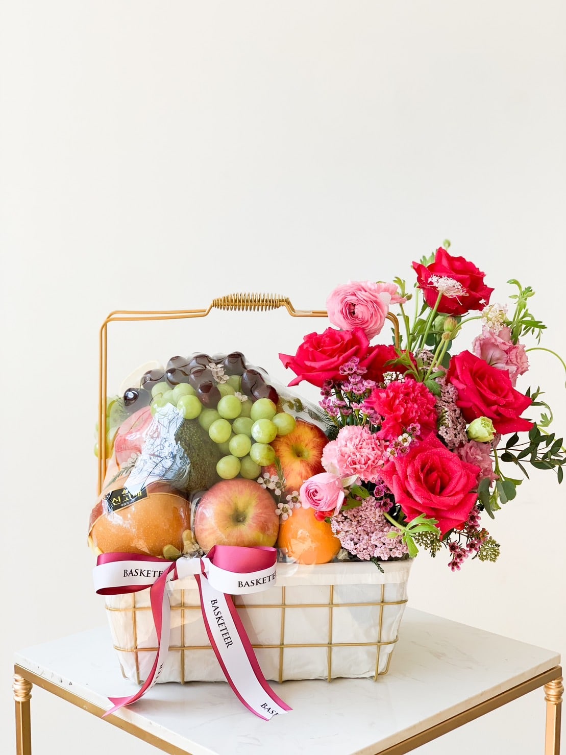 A white Fruit Rose Delight Basket filled with various fruits including apples, grapes, and oranges sits on a small table. A bouquet of vibrant pink and red flowers with green foliage is arranged on one side of the basket, accompanied by pink ribbons that read 
