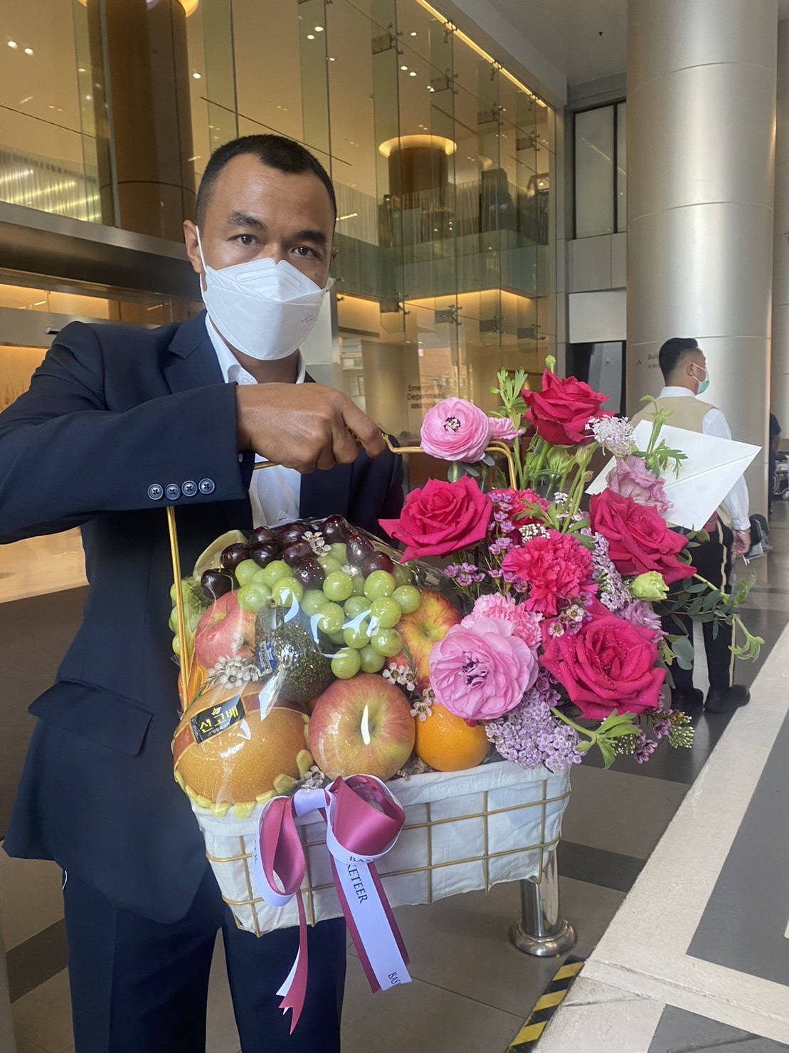 A man dressed in a suit and wearing a mask holds a large Fruit Rose Delight Basket filled with an assortment of fruits and vibrant flowers, including pink roses and ranunculus. The background shows a modern building lobby with glass and metallic accents.