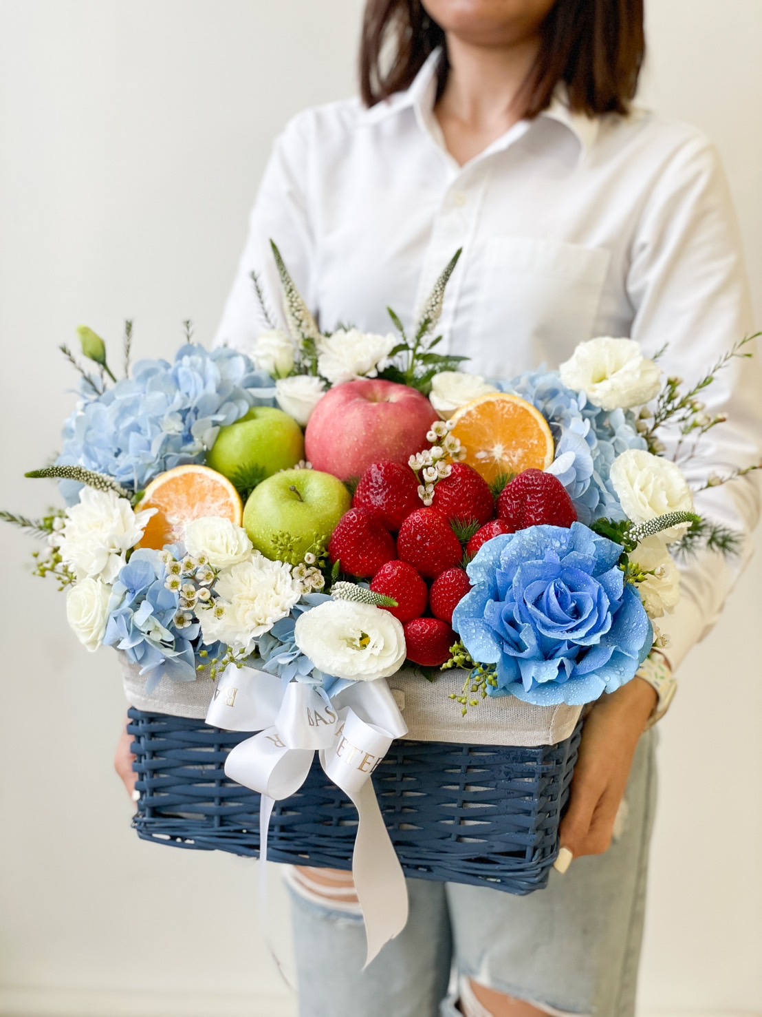A person in a white shirt holds a blue wicker basket filled with an assortment of colorful fruits and flowers, including apples, strawberries, blue hydrangeas, and white carnations. The basket is adorned with a white ribbon and a few sprigs of purple flowers for that Deluxe Fruit Basket touch.