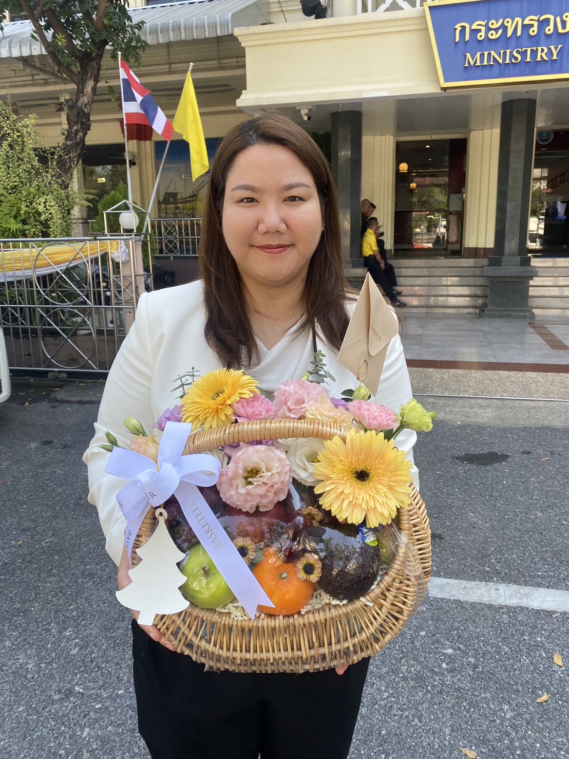 A woman holding a spring-themed fruit basket adorned with yellow gerbera daisies, pink flowers, and lavender ribbon.