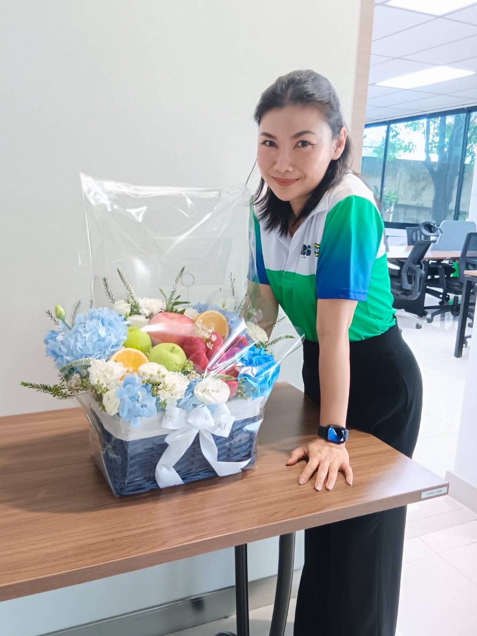 A woman wearing a green, white, and blue shirt stands next to a table with a Deluxe Fruit Basket containing an assortment of fruits and purple flowers, wrapped in transparent plastic. She is smiling and leaning slightly towards the basket. In the background is a bright office space.