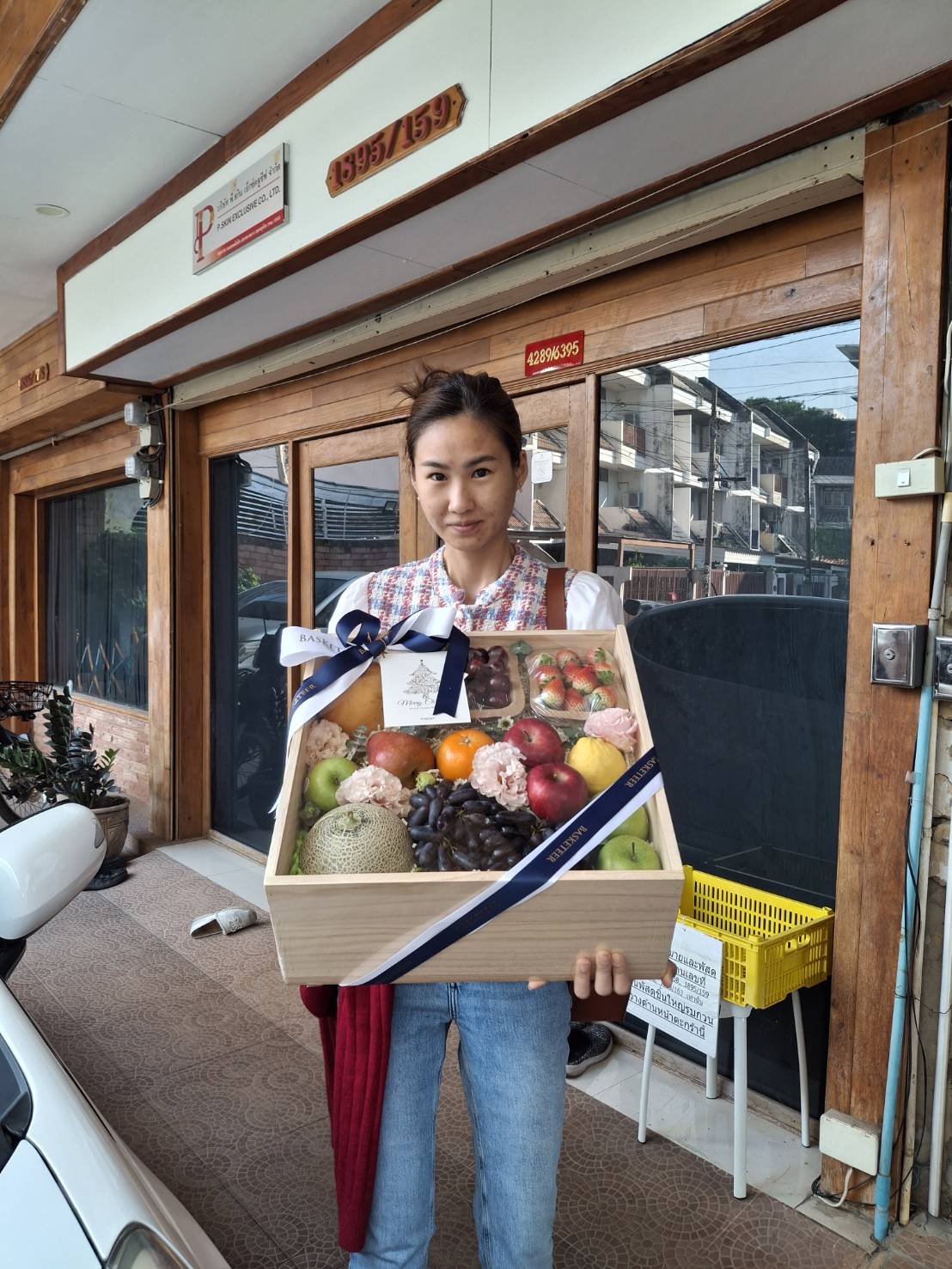 Premium wooden gift box filled with assorted fresh fruits, decorated with carnations and tied with a navy ribbon, held by a smiling recipient.
