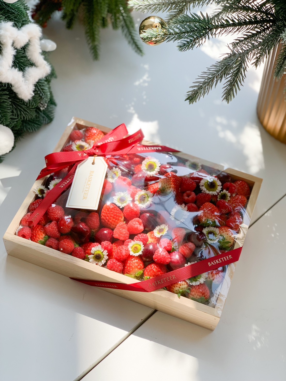 A wooden gift box filled with fresh strawberries, raspberries, cherries, and blueberries, accented with floral decorations and tied with a red ribbon.