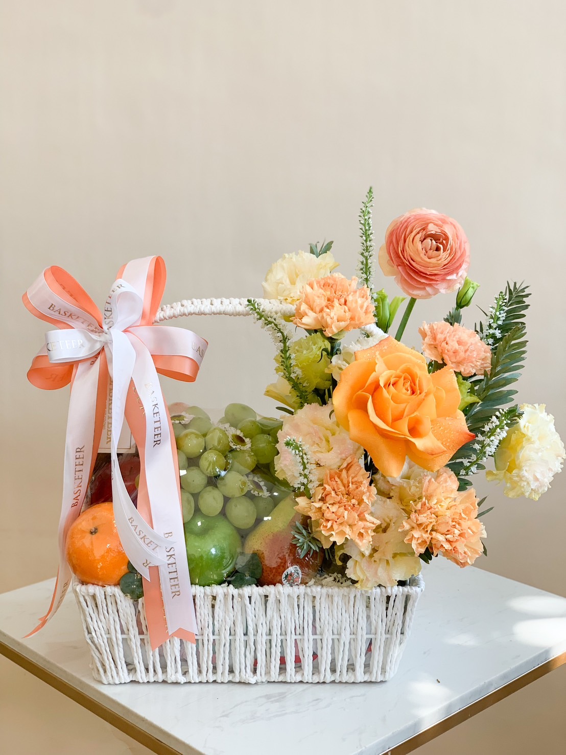 A white wicker basket filled with fresh fruits, decorated with orange roses, carnations, and pastel flowers, tied with an orange ribbon.