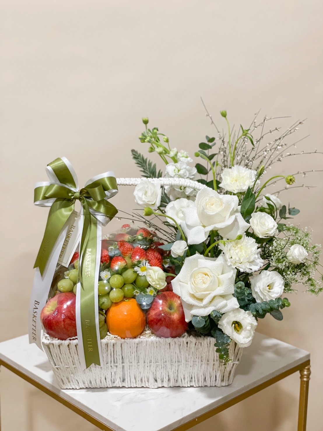 White basket with fresh fruits, including apples, oranges, strawberries, and grapes, paired with elegant white flowers by Basketeer.
