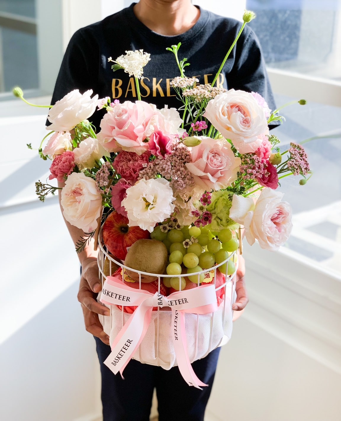 A person is holding a white basket filled with a mix of pink and white flowers, including roses and carnations. The Purple Blooms Fruit Basket also contains assorted fruits like grapes, a kiwi, and a pomegranate. Two pink ribbons with text 