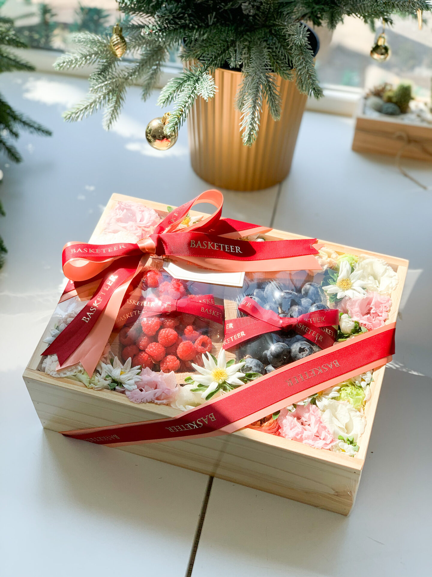 A wooden gift box containing fresh raspberries, blueberries, and a beautiful arrangement of pink and white flowers, tied with a red Basketeer ribbon.