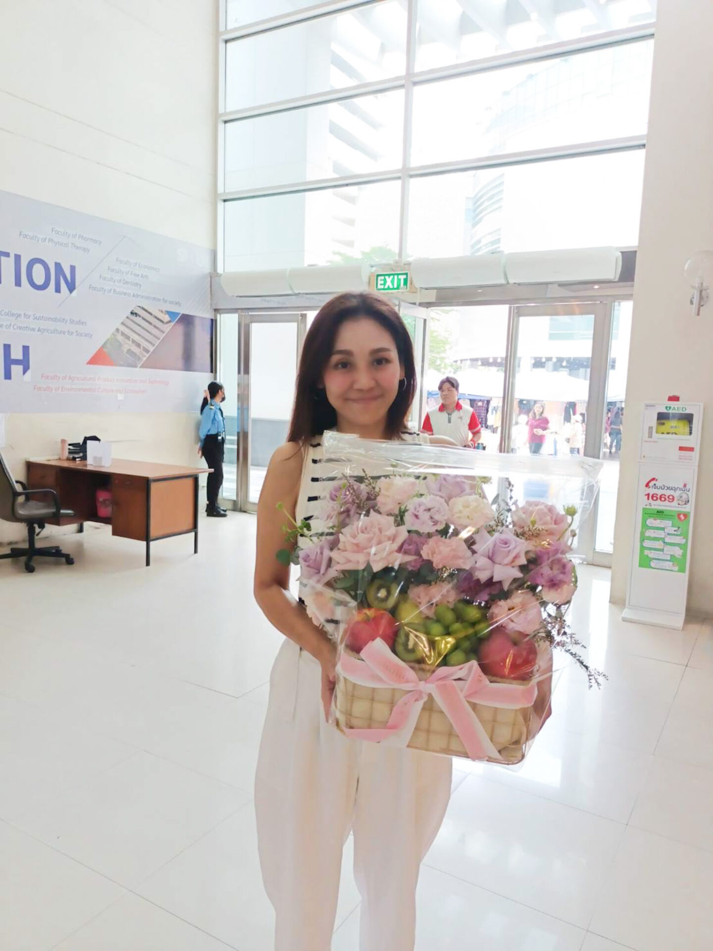 A woman in a white outfit stands in a bright lobby holding a Purple Blooms Fruit Basket wrapped in clear plastic and tied with a pink ribbon. Behind her is a reception desk on the left and glass doors leading outside in the background.