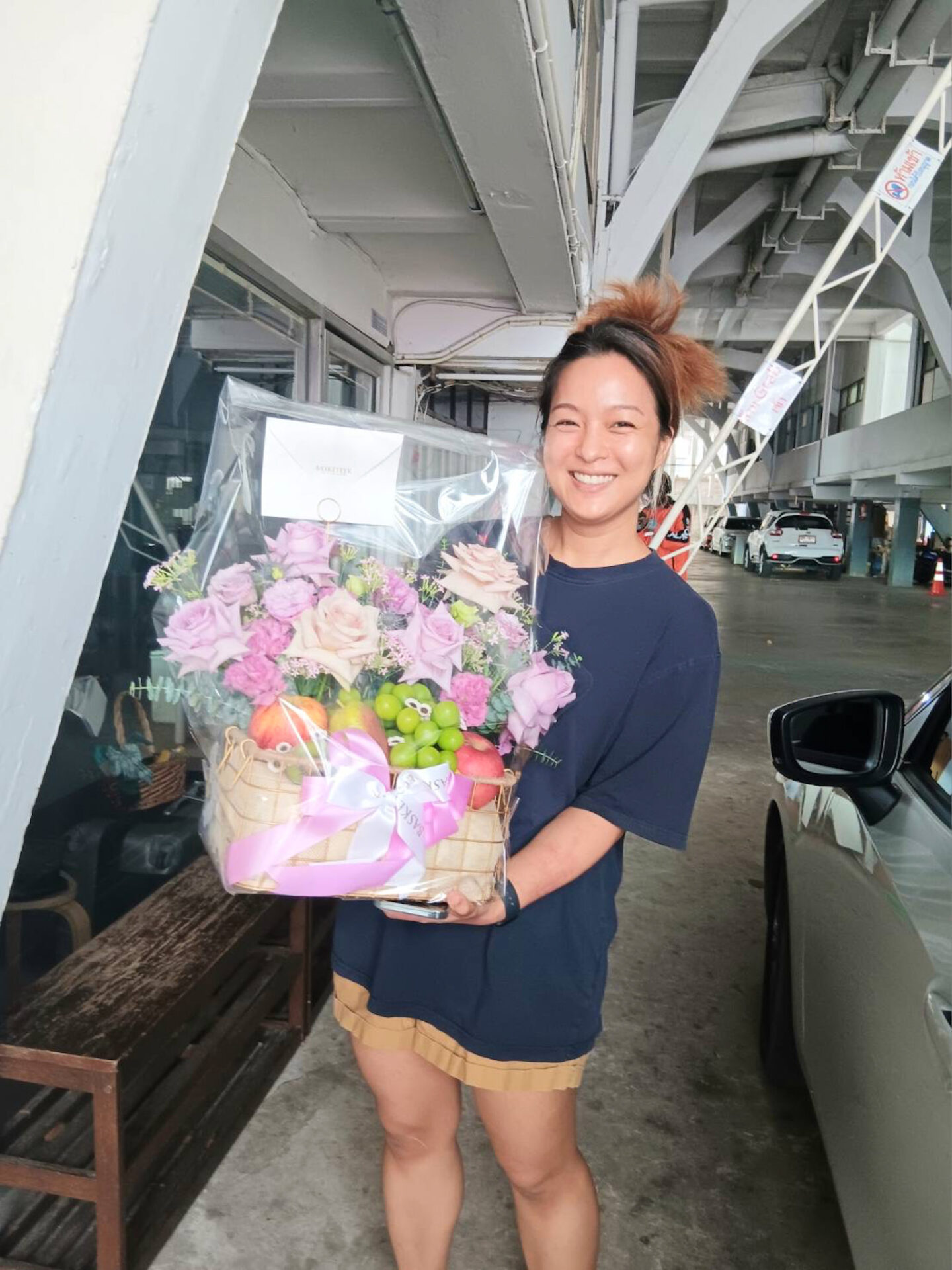 A smiling person stands indoors holding a basket filled with purple blooms and green apples wrapped in cellophane with a ribbon. They are wearing a navy blue t-shirt and beige shorts. A car and structural beams are visible in the background.