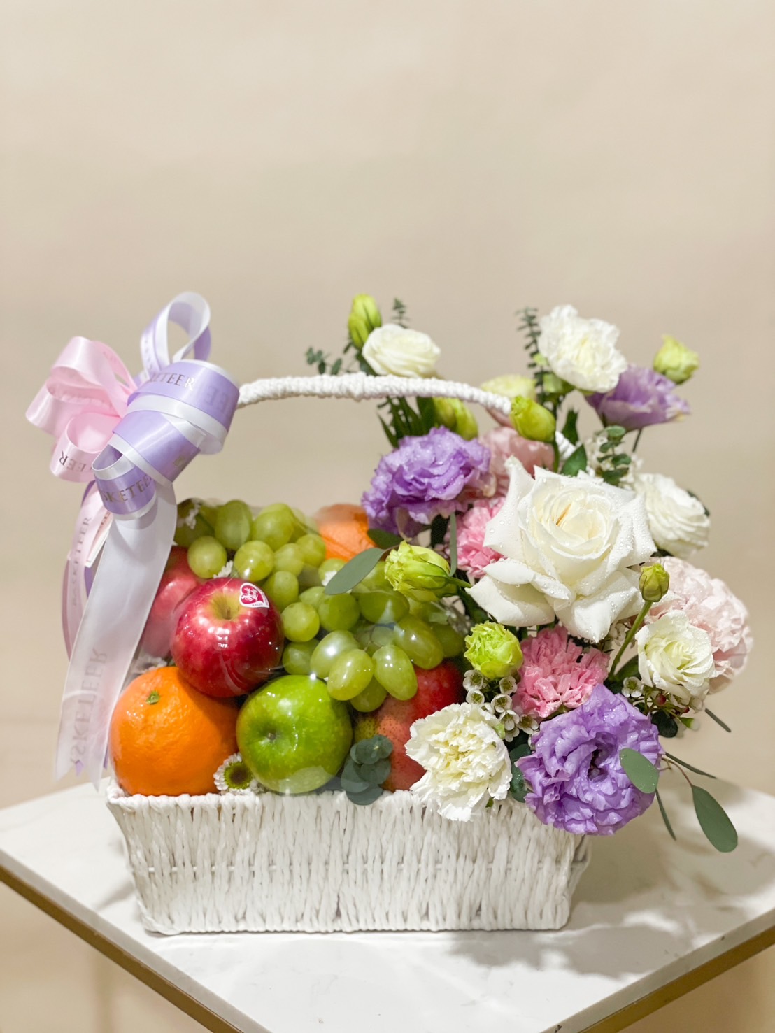 A white woven basket filled with fresh fruits and a vibrant flower arrangement of roses, carnations, and lisianthus, adorned with purple and pink ribbons.