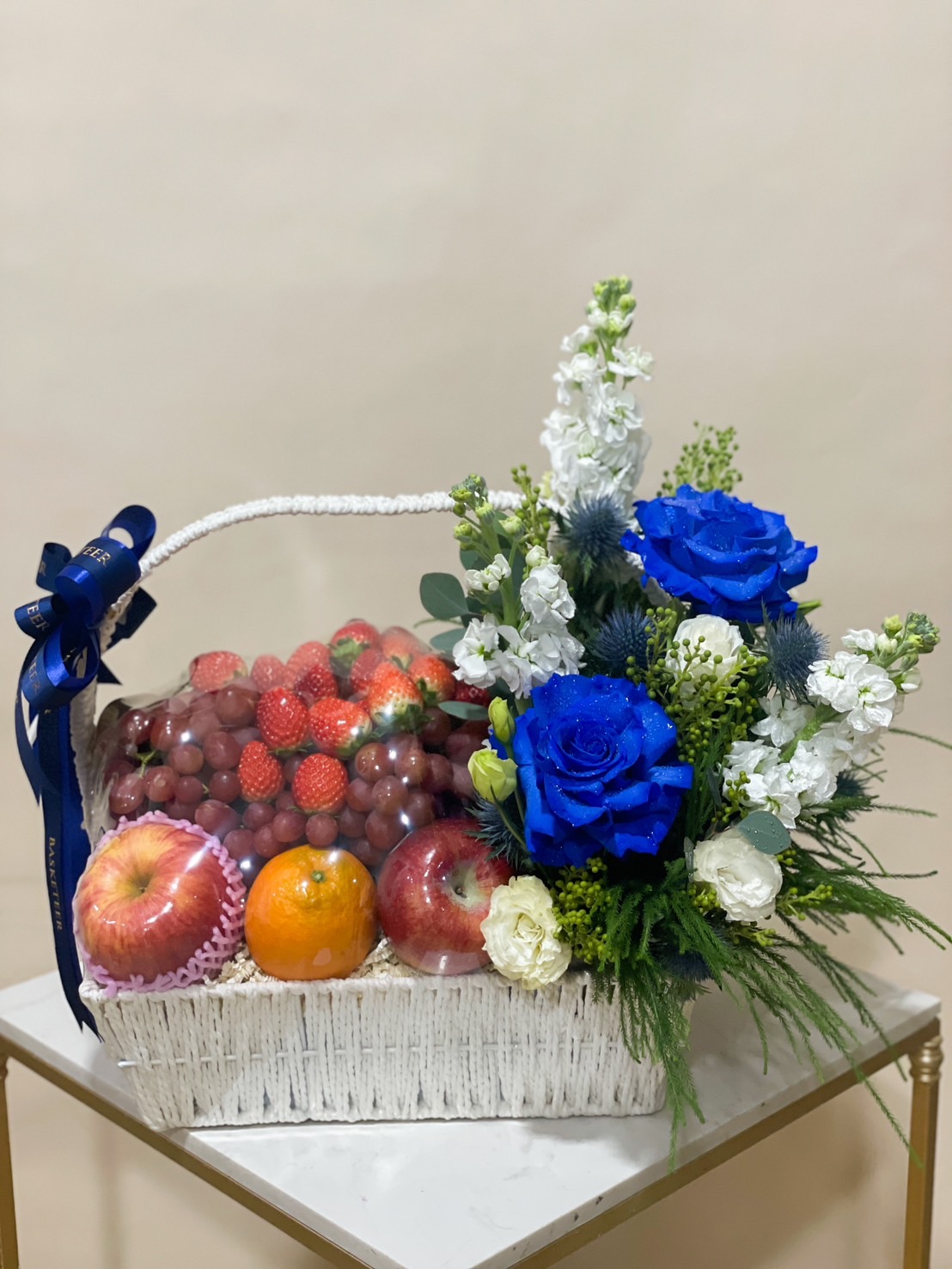 White woven basket filled with strawberries, red grapes, apples, and oranges, adorned with blue roses and white flowers.