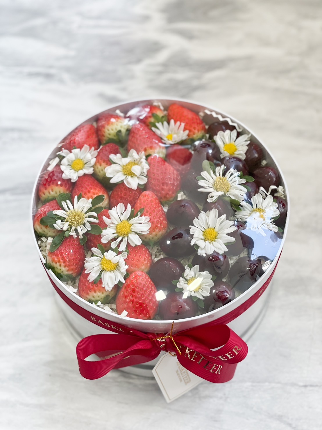 Round gift box filled with fresh strawberries, cherries, and daisies, adorned with a red ribbon.