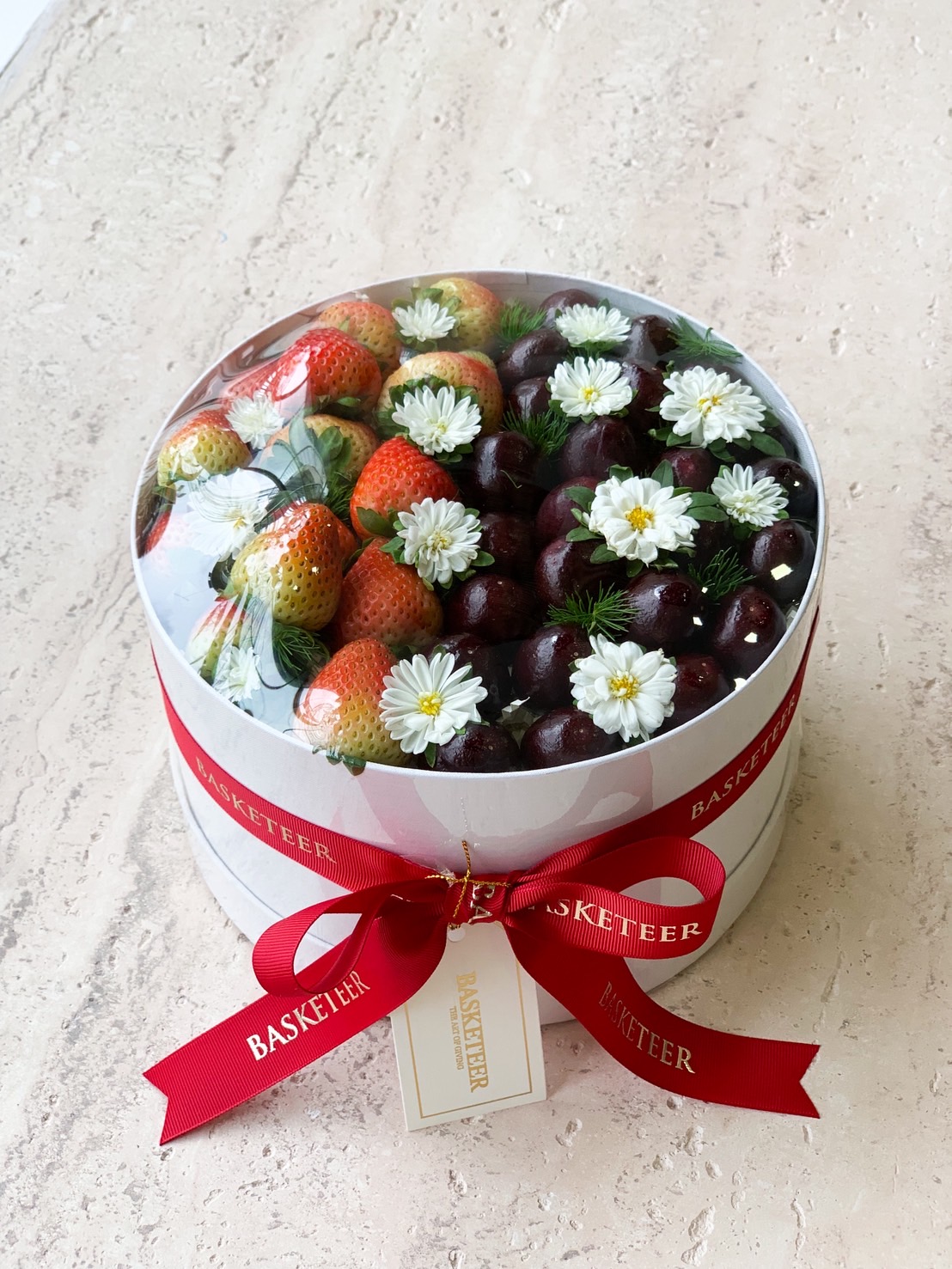 Luxury round fruit box with fresh strawberries, cherries, and white daisy flowers decorated with red ribbon