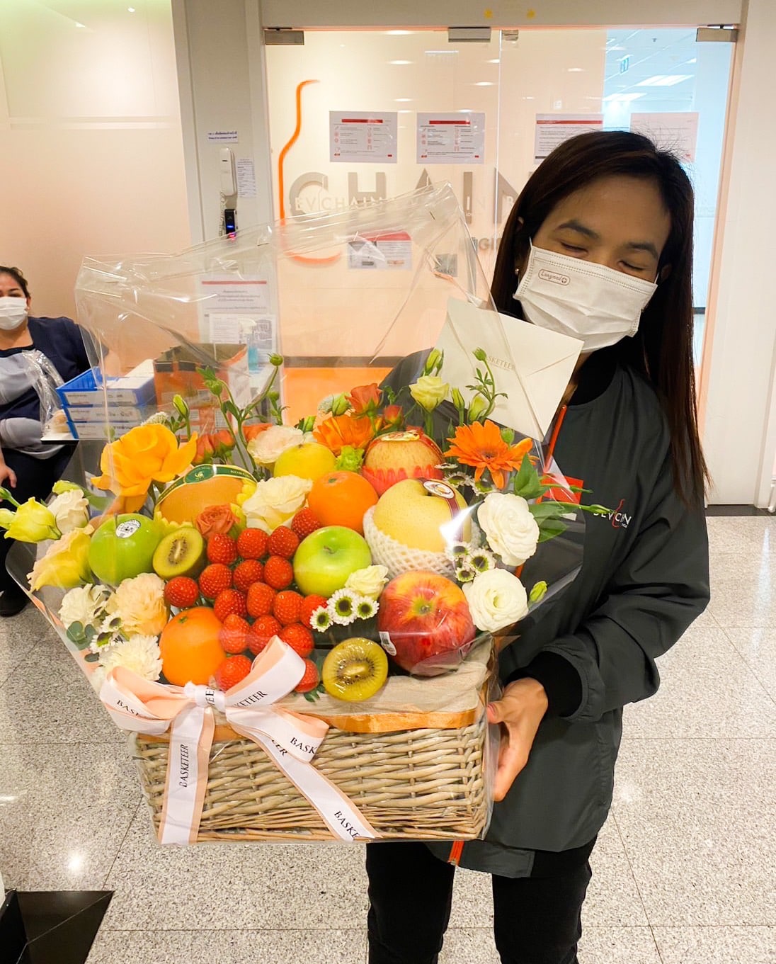 A person wearing a mask is holding a large mixed fruit flower basket filled with apples, oranges, kiwis, strawberries, and colorful blooms. They are standing indoors near an office reception area. Another masked person is seated in the background.
