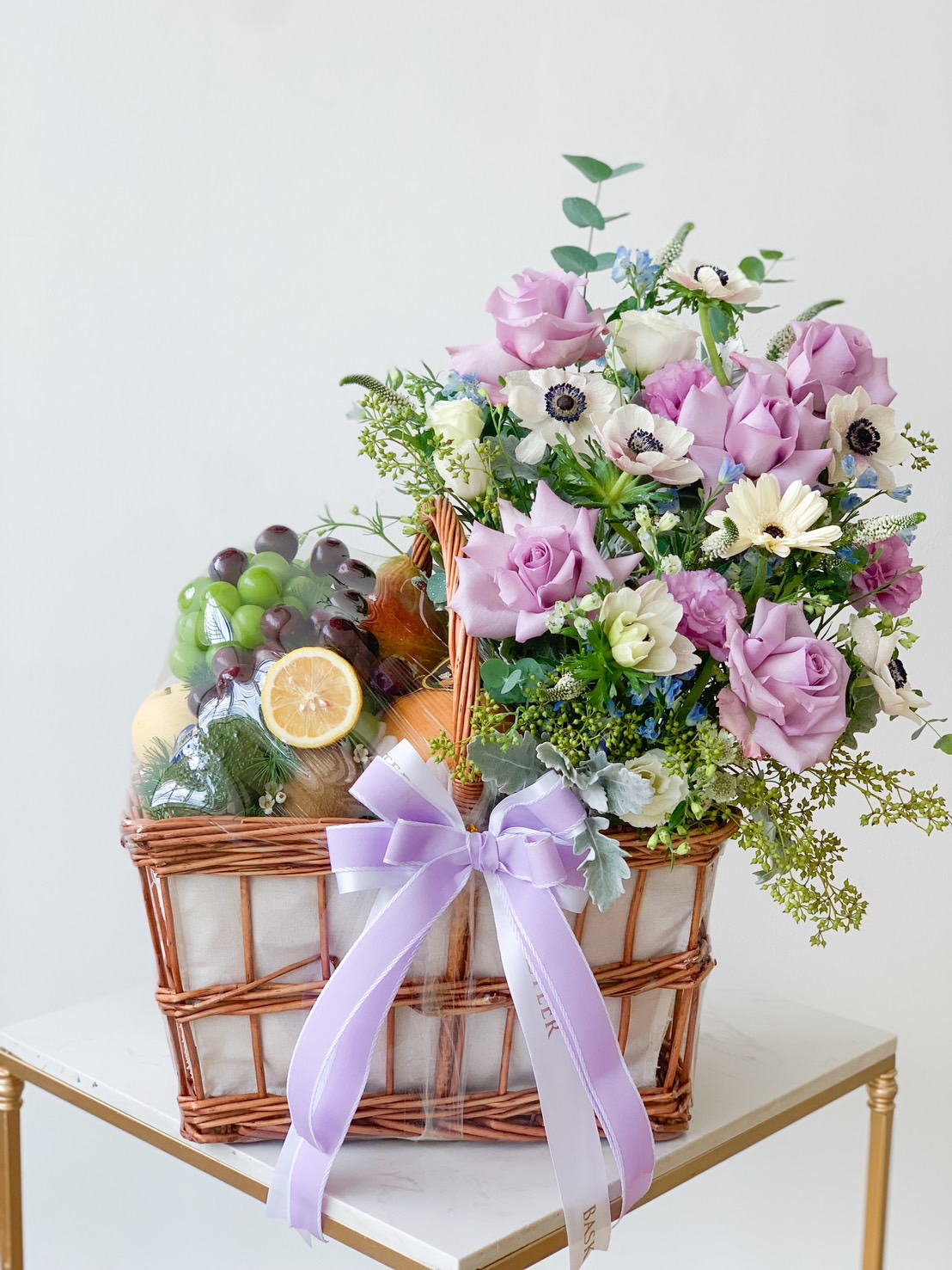 A wicker basket filled with fresh fruit and a colorful flower arrangement, featuring purple roses, white daisies, and greenery. The Fresh Fruit & Blue Bloom Basket is adorned with a light purple ribbon and is placed on a white table against a plain background.