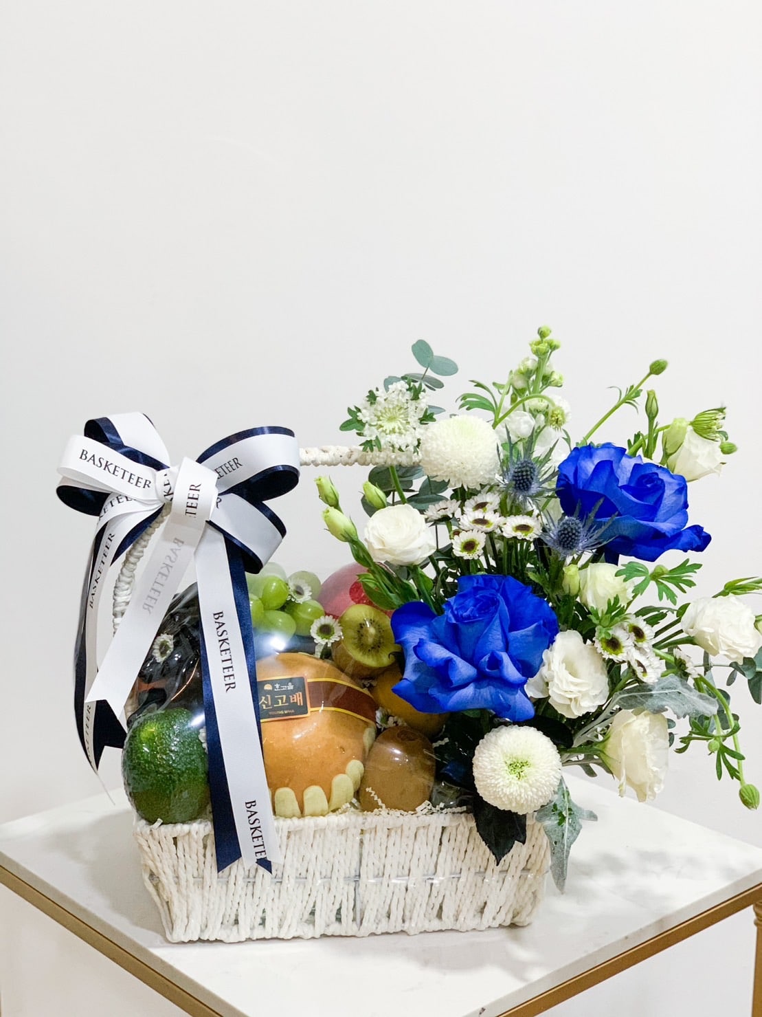 A white woven basket filled with various fresh fruits and a vibrant flower arrangement, showcasing blue roses and white blooms. The Blue Rose Fruit Basket is adorned with a white and black ribbon bow, with the handle also featuring a ribbon reading 