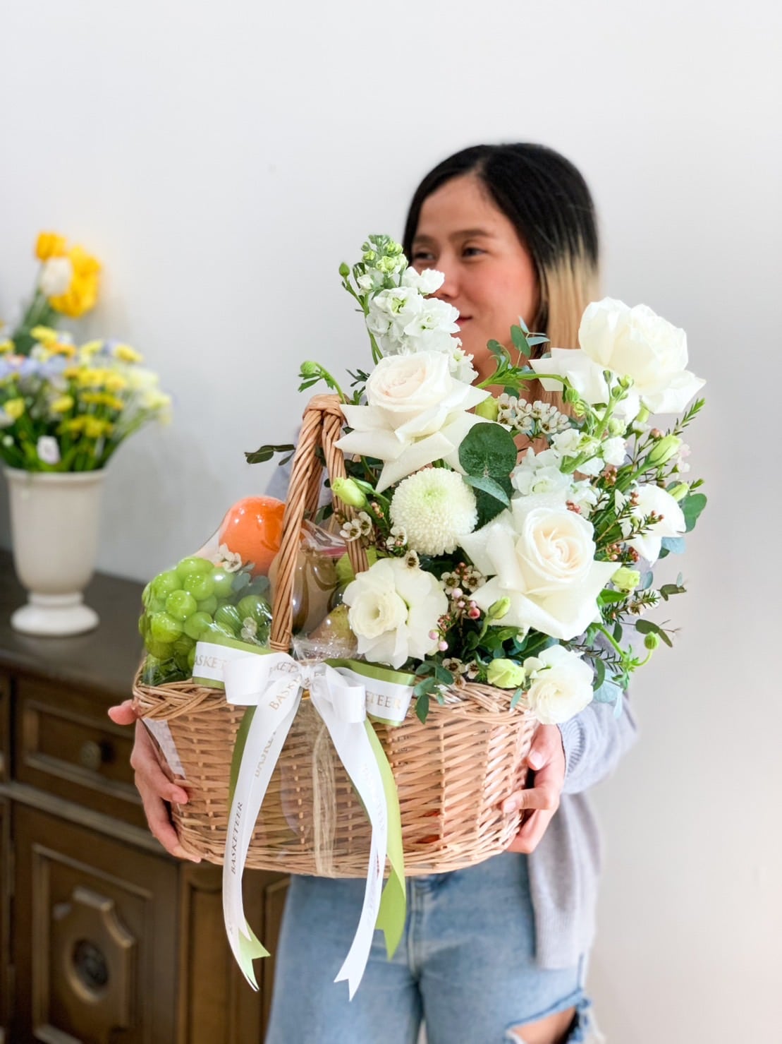 A person holds a wicker basket filled with white flowers, fresh fruit, and green grapes. The individual is smiling and wearing a light-colored shirt, with a vase of yellow flowers and greenery on a wooden cabinet in the background.