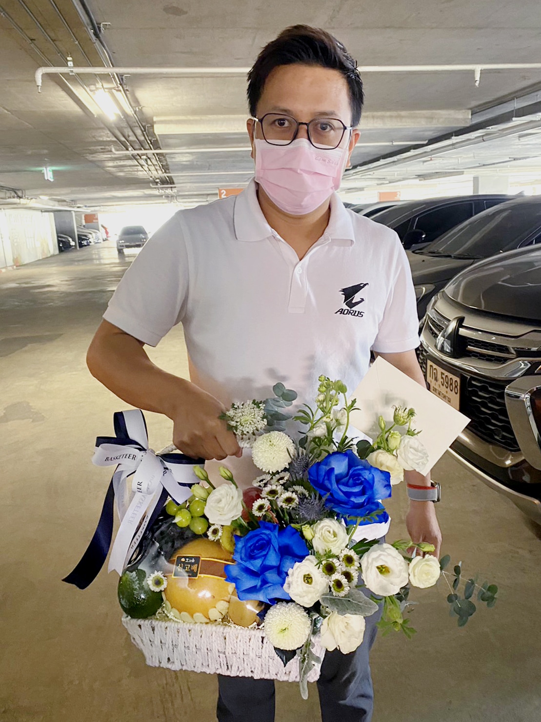 A man wearing glasses and a pink mask stands in a parking garage, holding a white fruit basket filled with blue roses, blue and white flowers, a yellow melon, and a leafy green fruit. He's dressed in a white polo shirt with a black emblem and wears an elegant wristwatch.