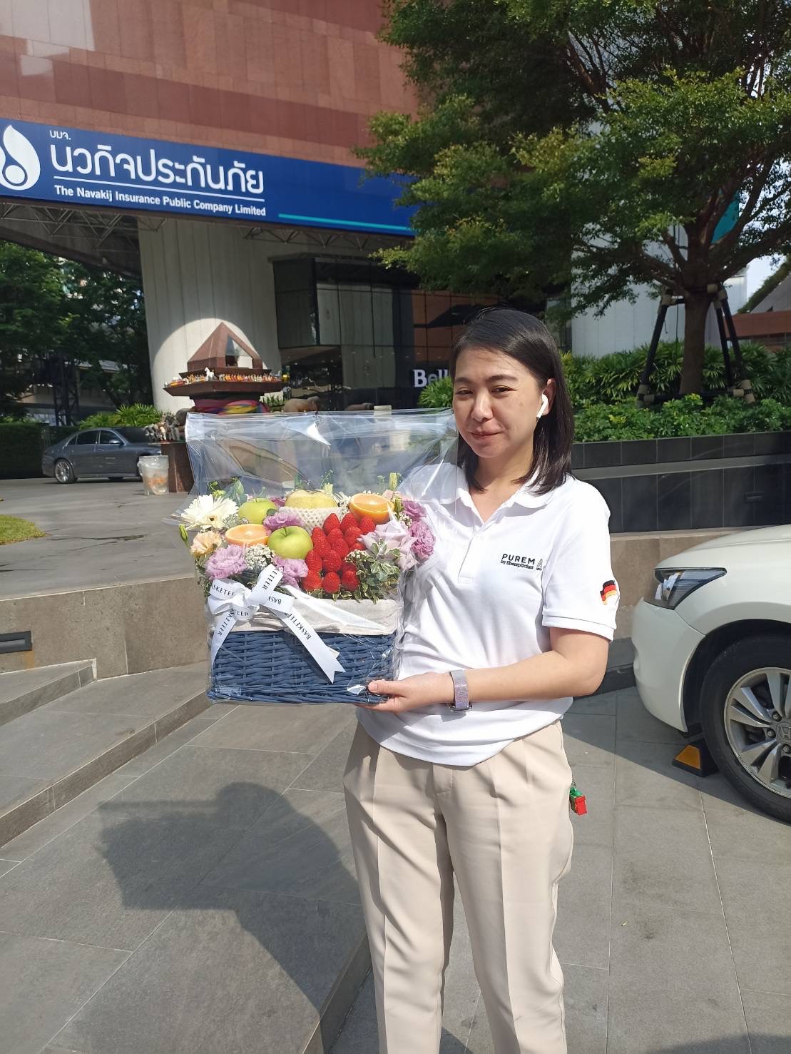A woman wearing a white polo shirt with a logo on the sleeve holds a bouquet of colorful flowers and fruits. She is standing on a sidewalk with a building and a sign for 