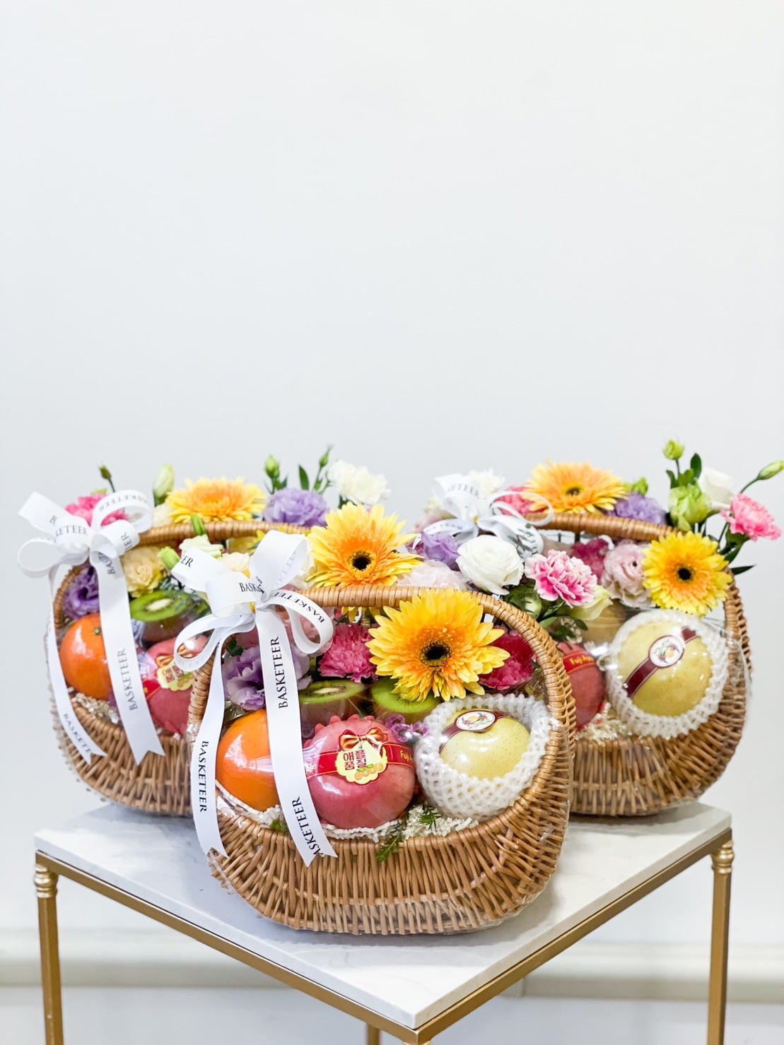 Three Luscious Fruit Harvest Baskets filled with a variety of colorful flowers, including yellow and purple blooms, and fresh fruit like apples and oranges are arranged on a small table. Each basket is adorned with white ribbons that have text printed on them.