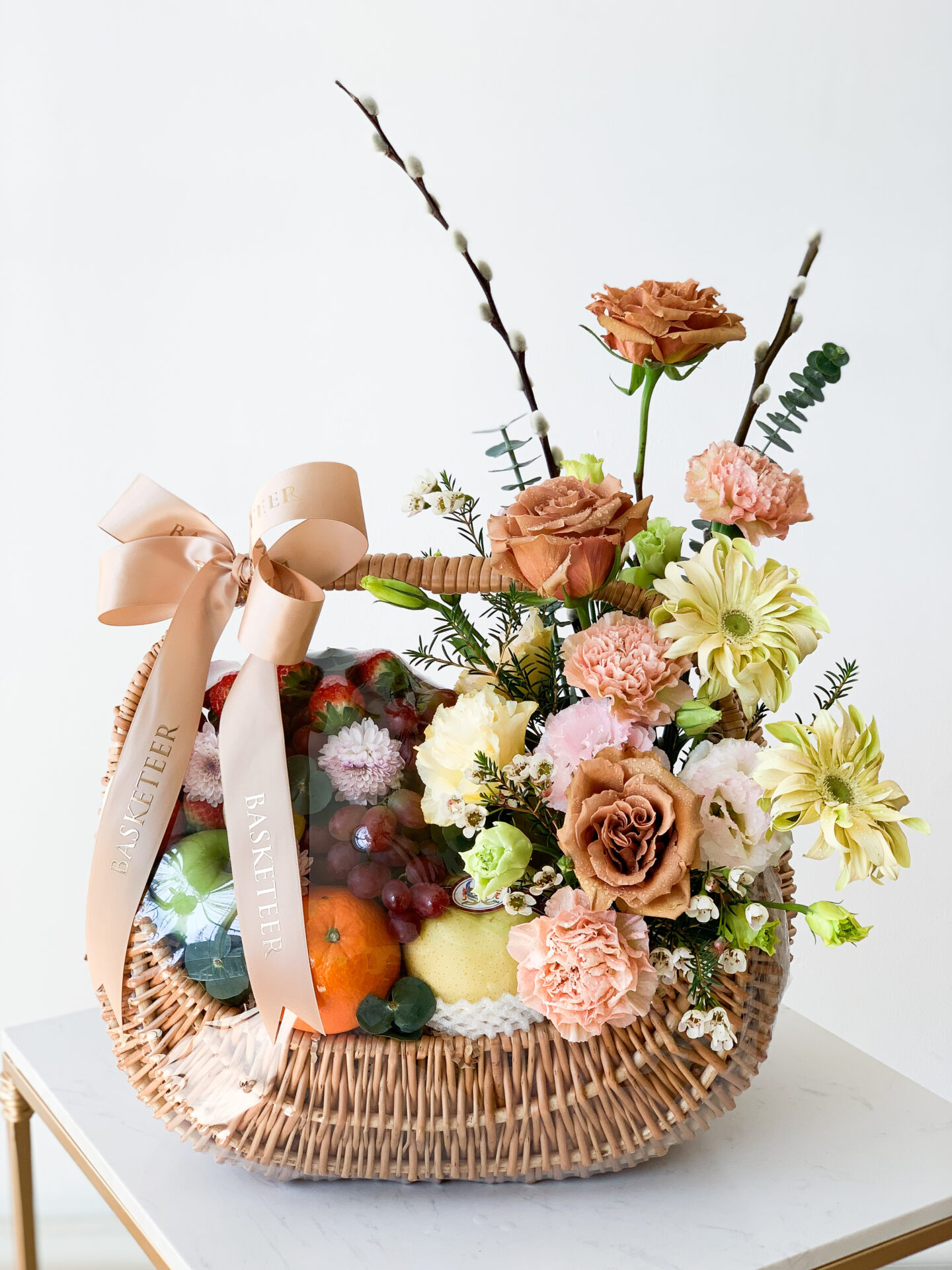 A wicker basket filled with a mixed arrangement of fresh fruit, including oranges, grapes, and other delights, accompanied by a diverse bouquet of flowers such as roses, carnations, and lilies. Adorned with a large beige ribbon bow marked 