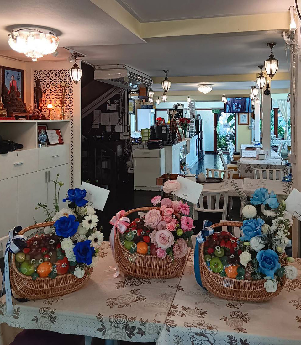 A cozy, well-lit interior of a café or restaurant with tables adorned with three wicker baskets. Each basket contains a colorful arrangement of fresh fruit and vibrant toffee flowers, including blue, pink, and white blooms. The ambiance is inviting and homely.