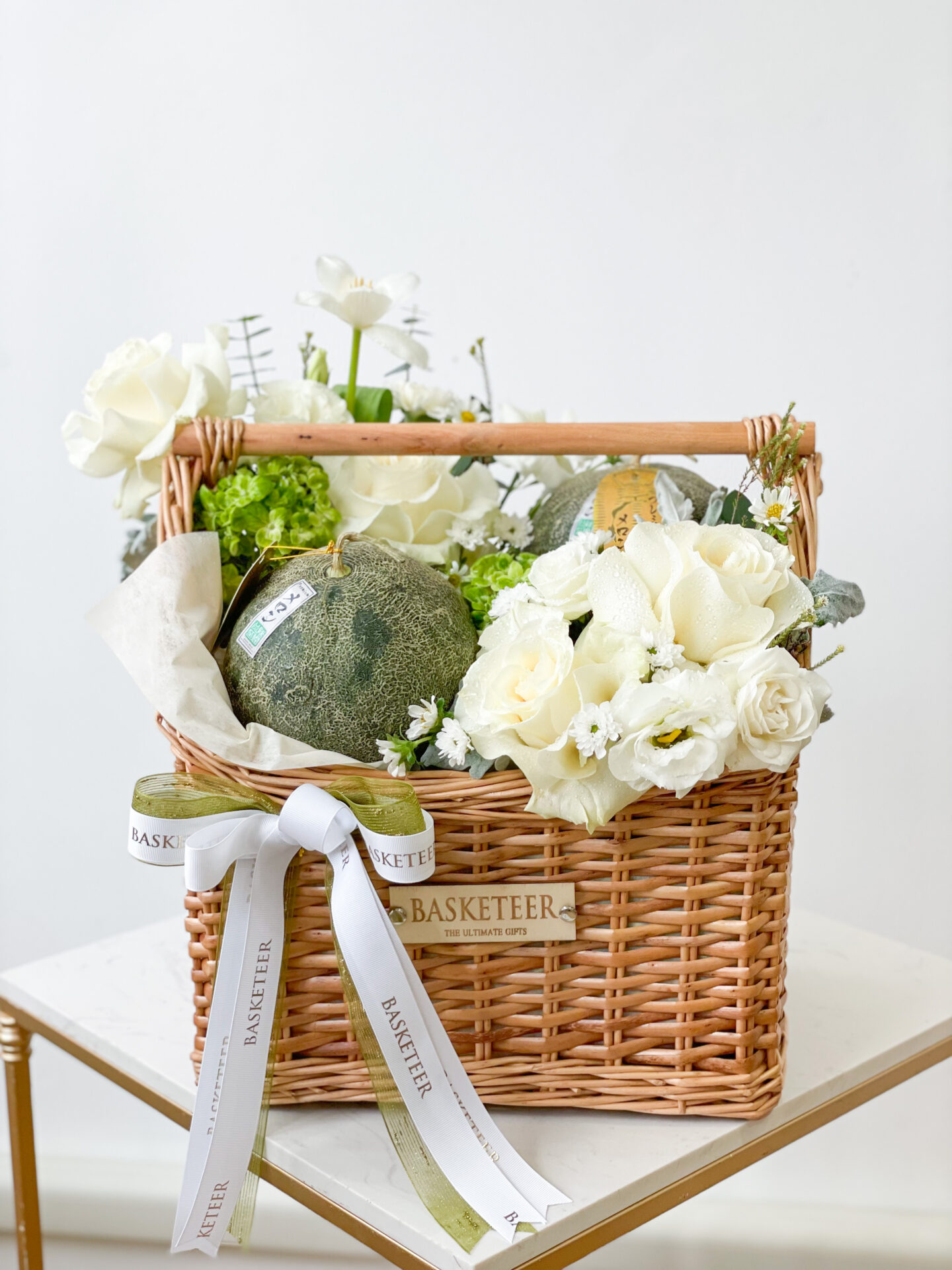 A wicker basket filled with a variety of white flowers and greenery, adorned with a white ribbon imprinted with 