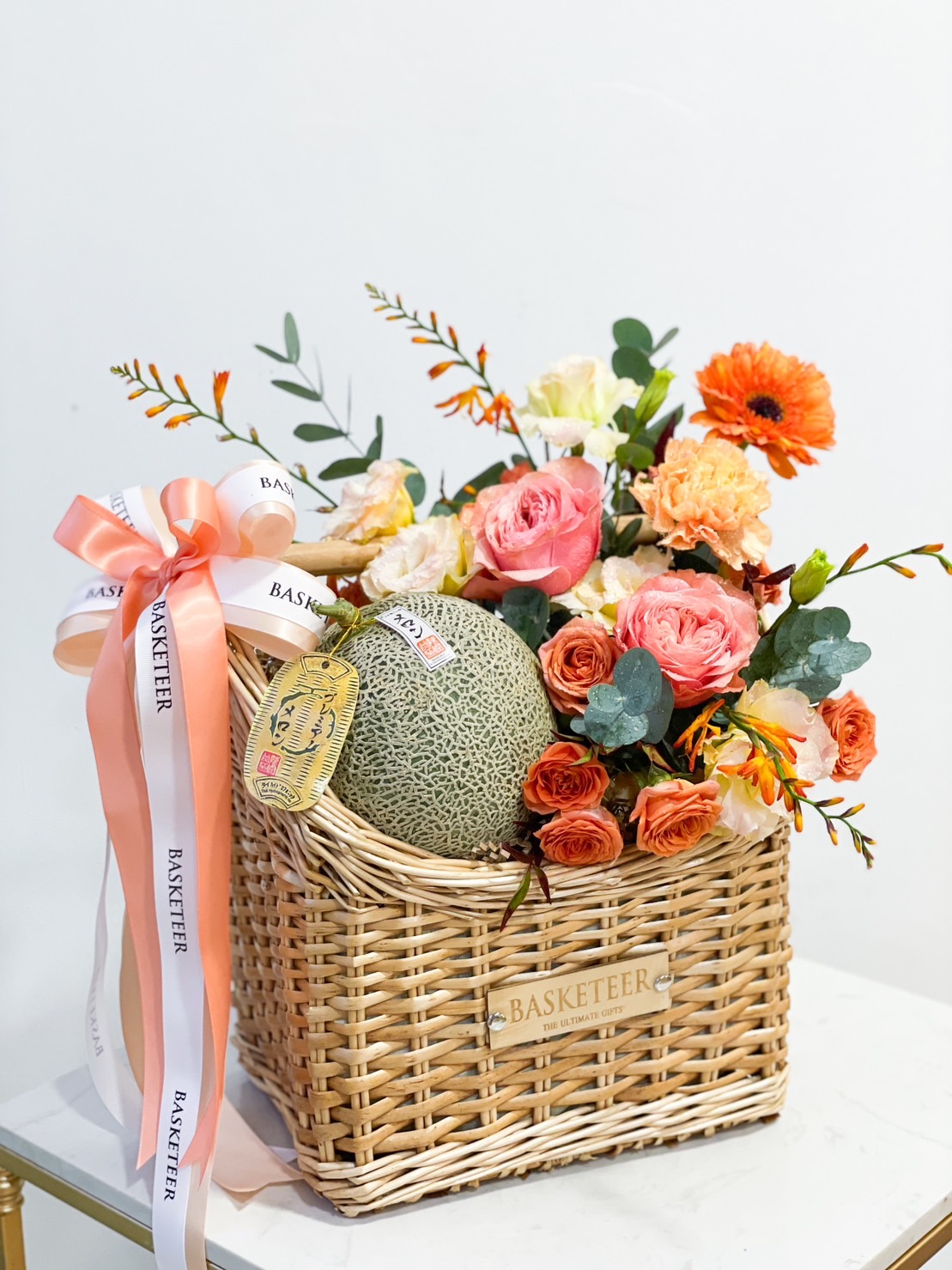 A wicker basket filled with assorted orange, pink, and white flowers, green foliage, and a cantaloupe. The ribbon features the word 