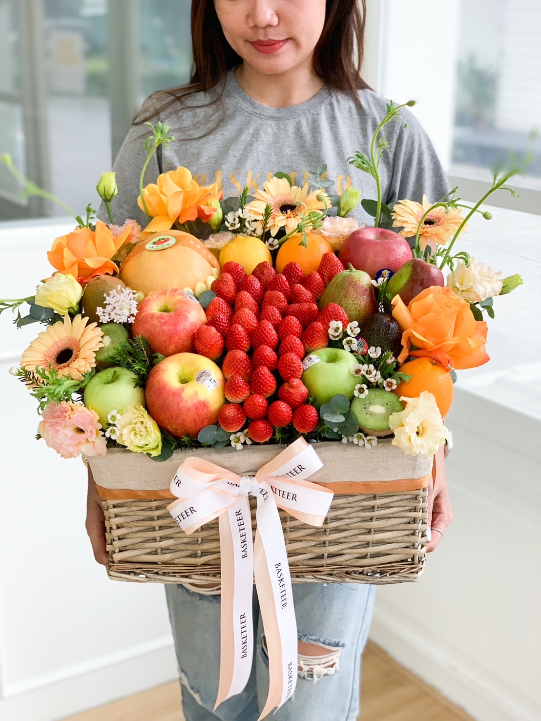 A person holding a wicker basket filled with an assortment of colorful fruits, including apples, strawberries, grapes, and mangoes. The blooming fruit gift basket is also adorned with vibrant flowers such as roses and gerberas and is tied with a pink ribbon that says 