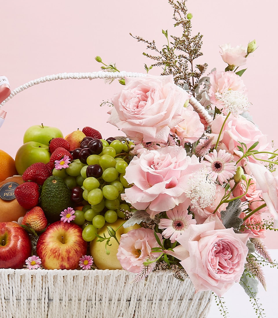A luxurious white wicker basket, elegantly filled with an assortment of premium fruits such as crisp apples, succulent grapes, and vibrant oranges, is opulently adorned with delicate pink roses and dainty white blossoms interwoven with lush green foliage. The basket's refined presentation showcases the Basketeer brand ribbon subtly draped across its handle. Set against a soft pink backdrop, this breathtaking floral and fruit ensemble invites indulgence and celebrates opulence.