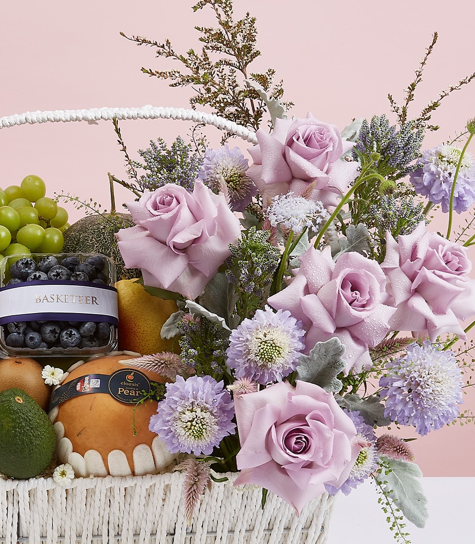 Presenting the Basketeer Radiant Fruits & Purple Blooms Basket: An exquisite white wicker basket brimming with ethereal light purple roses and delicate lavender-colored flowers interwoven with lush green foliage. Nestled among this botanical splendor is a carefully curated selection of sumptuous fruits—crisp green grapes, plump blueberries, succulent pears, and a rich avocado—all arranged against an inviting light pink backdrop. The subtle elegance of a ribbon adorned with the Basketeer logo enhances the luxurious presentation, making this basket an irresistible centerpiece for any occasion.