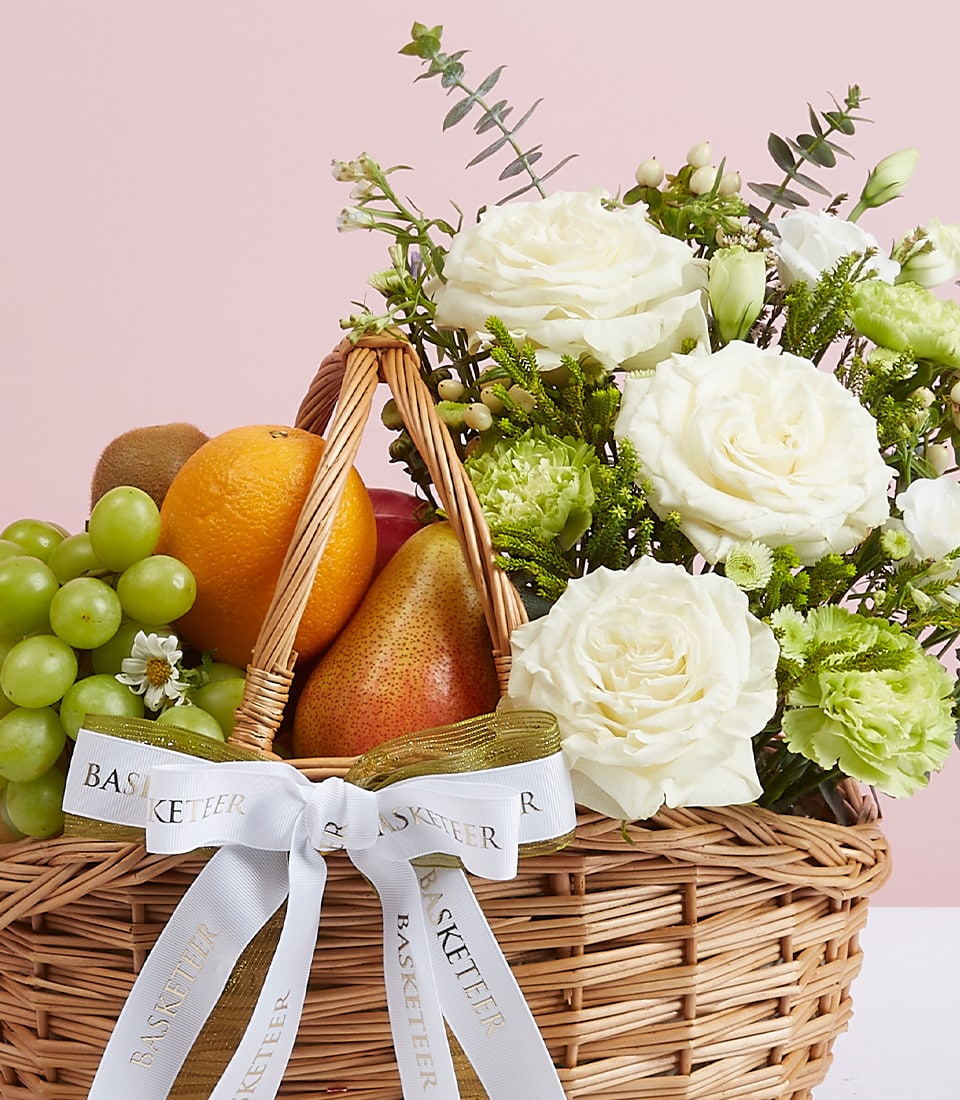 A lavish wicker basket brimming with an assortment of vibrant fresh fruit—succulent green grapes, luscious oranges, crisp apples, and juicy pears—alongside an elegant bouquet of pristine white roses adorned with lush greenery. The arrangement is beautifully presented with a white satin ribbon bearing the distinguished 