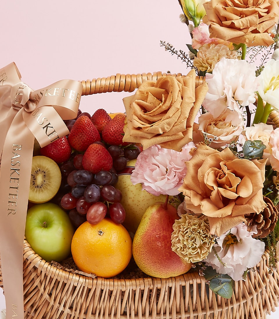 A luxurious woven basket brimming with an array of fresh fruits, including crisp green apples, succulent strawberries, juicy grapes, a ripe pear, and a vibrant orange. The bounty is elegantly adorned with exquisite tan and pink roses, delicate white flowers, and a sophisticated beige ribbon. The Basketeer logo graces the scene, adding an extra touch of elegance to this inviting display.
