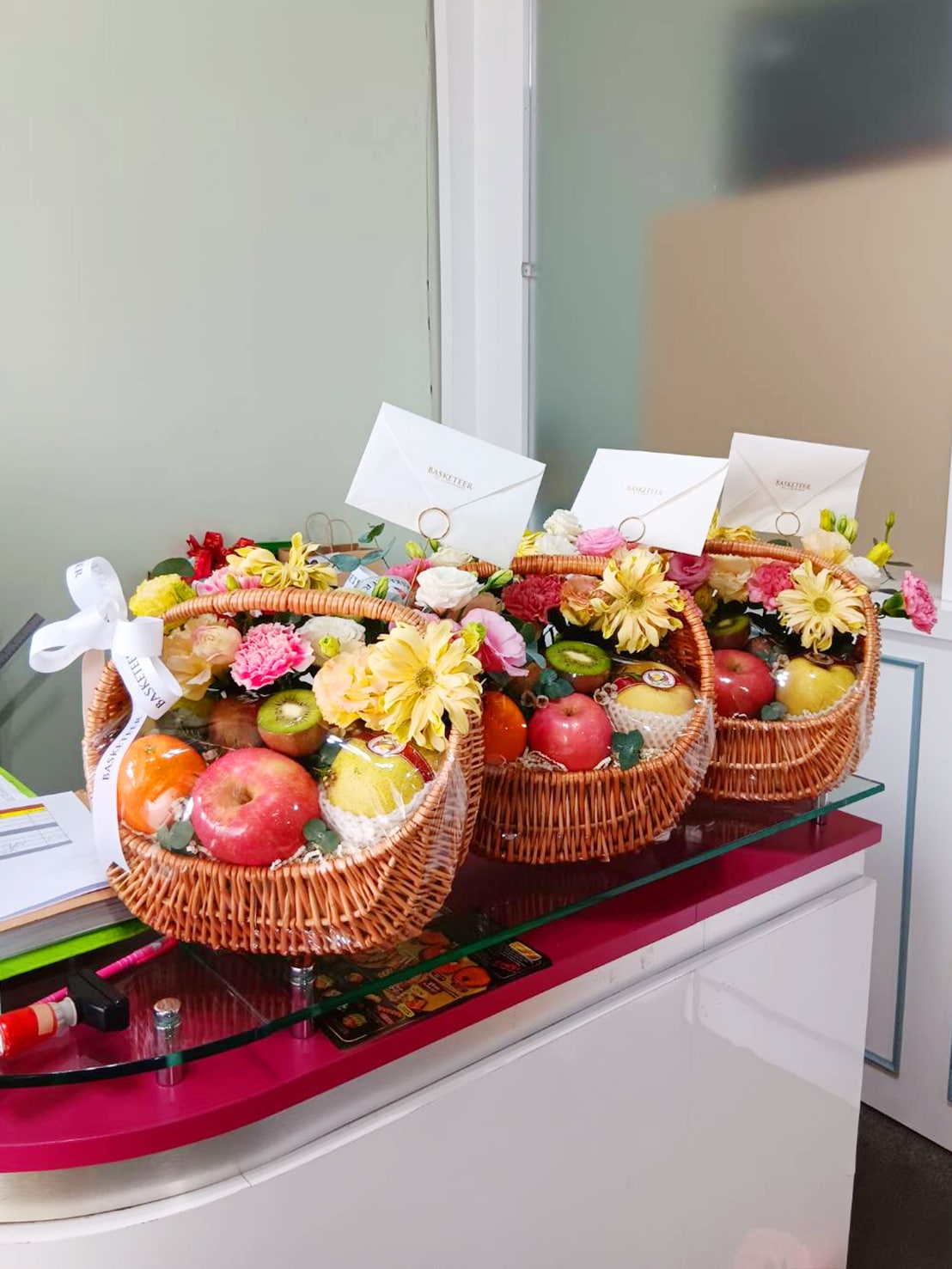 Three decorated baskets of assorted fruits, including apples, oranges, and kiwis, showcased on a glass table. Each Luscious Fruit Harvest Basket is adorned with fresh flowers and white envelopes, featuring a ribbon with the word 